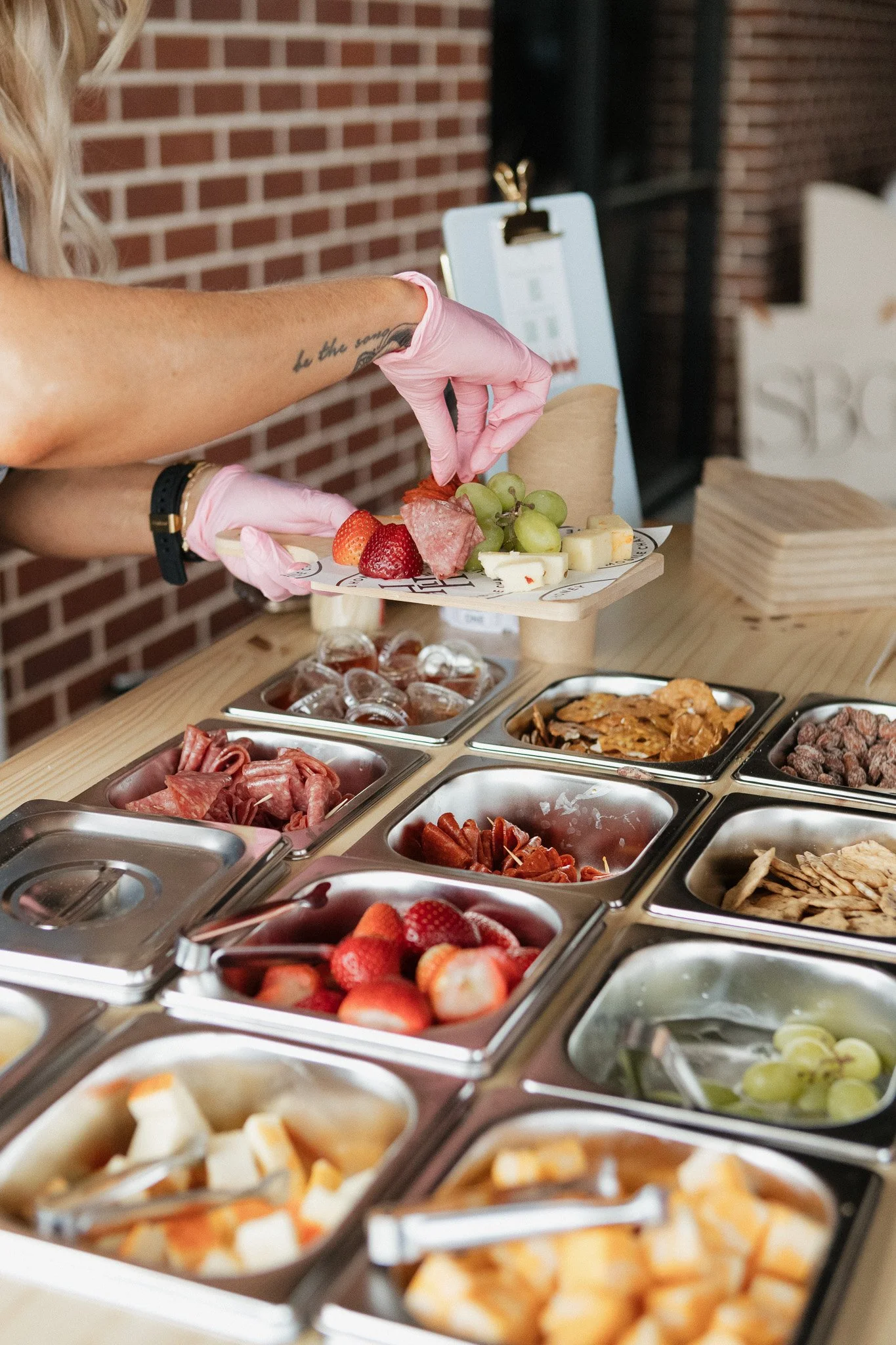 A person wearing pink gloves placing strawberries and grapes on a cheese platter at a buffet table. Various trays of sliced fruits, meats, cheeses, and snacks are arranged on the table.