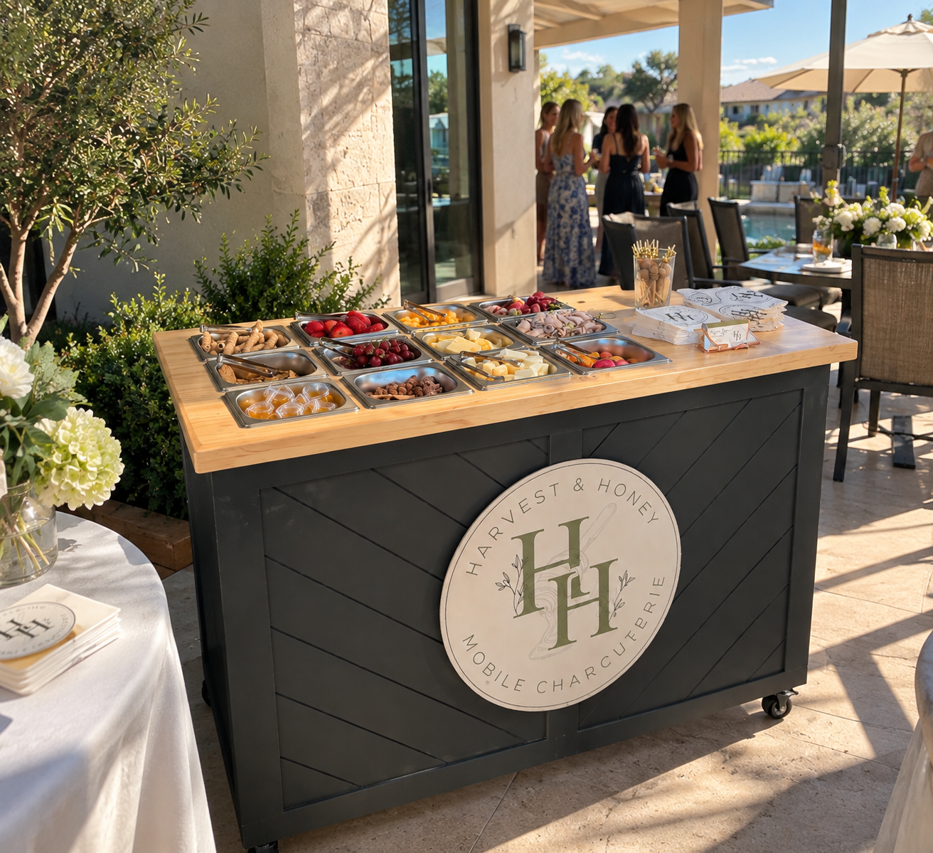 A buffet table with various fruits, cheeses, and snacks set outdoors next to a pool with a group of women socializing in the background.