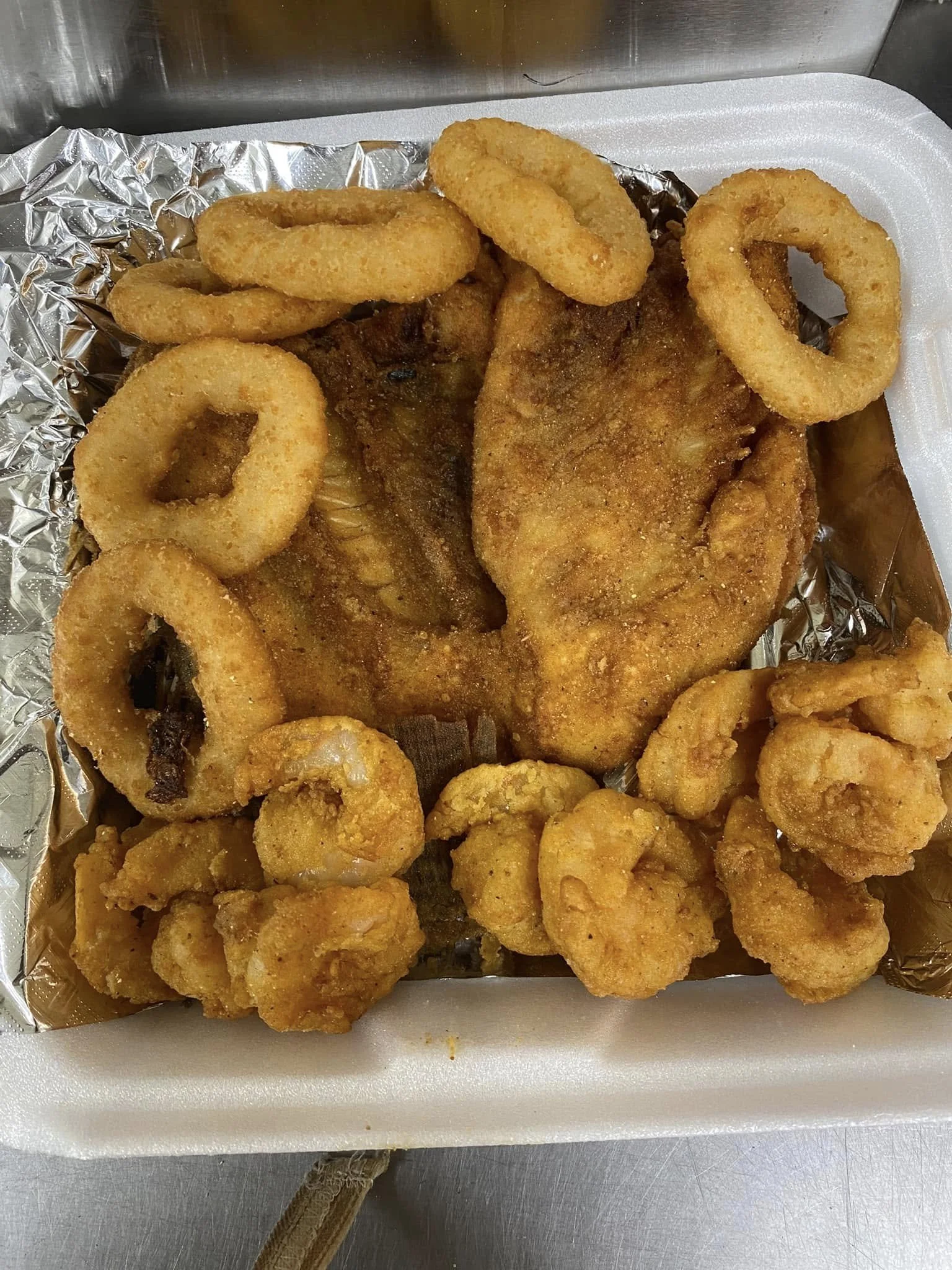 Fried fish fillet with breaded onion rings and fried shrimp on a foam tray lined with aluminum foil.