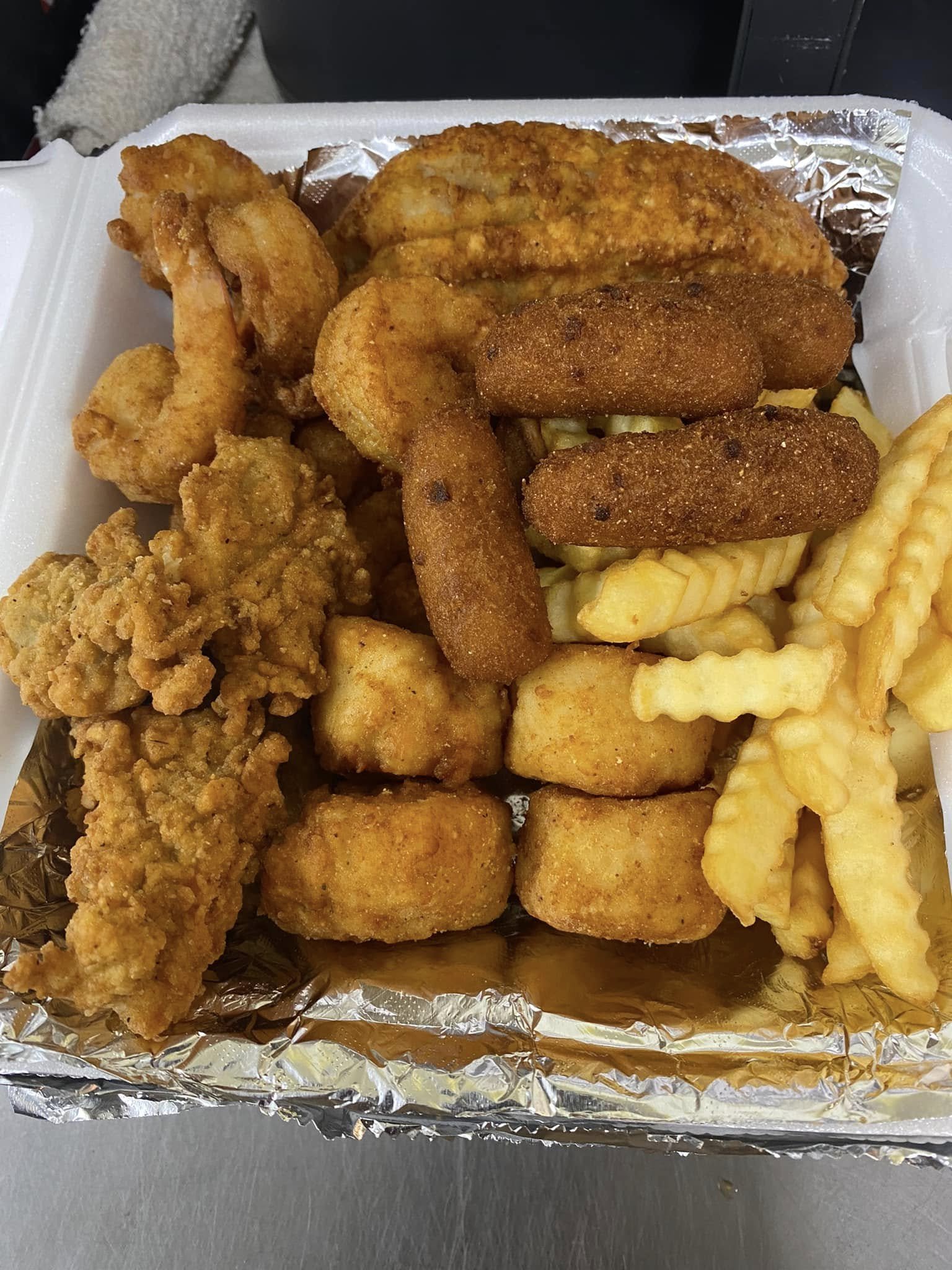 A takeout tray filled with fried chicken tenders, breaded mozzarella sticks, French fries, and what appears to be fried chicken pieces, all lined with aluminum foil inside a foam container.