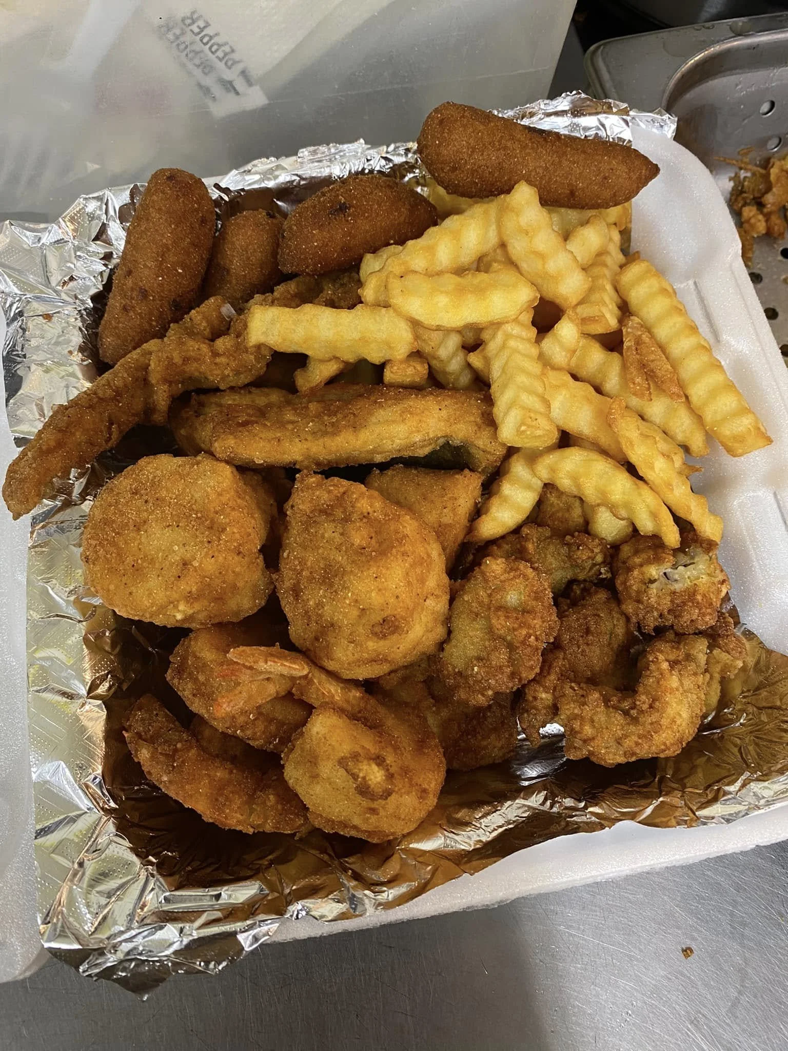 Fried chicken, French fries, and fried fish pieces in a styrofoam container lined with aluminum foil.