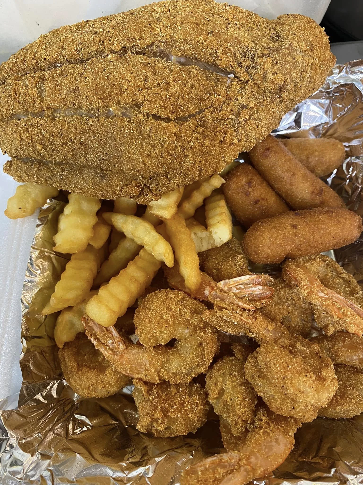 Close-up of fried chicken tenders with a golden-brown crispy coating, crinkle-cut French fries, and fried shrimp on aluminum foil.