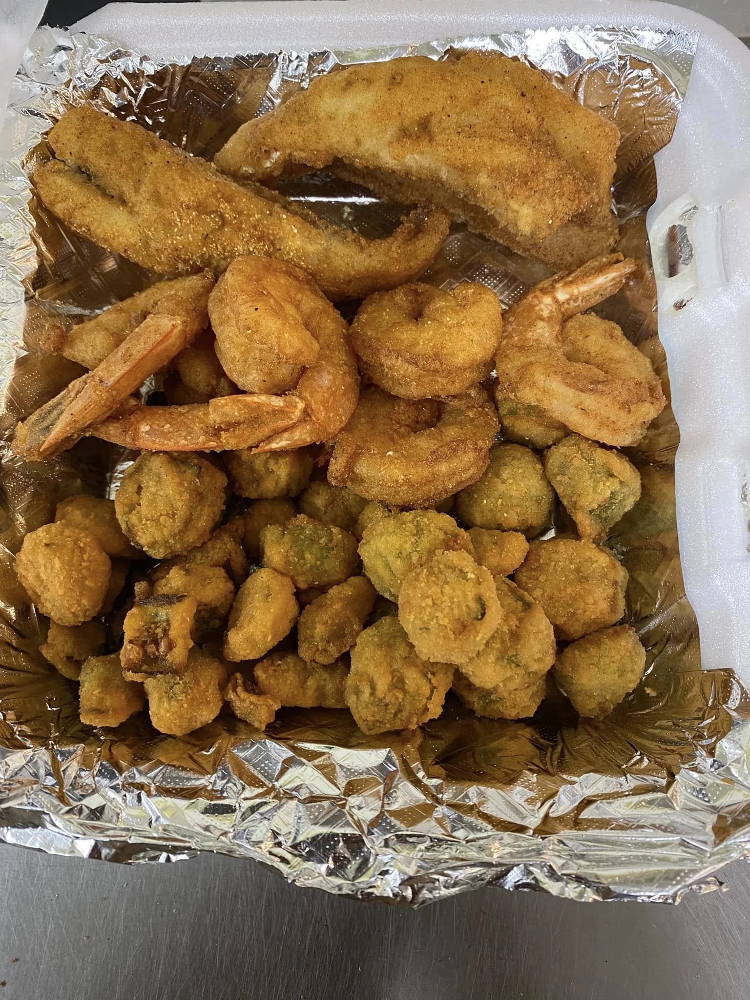 A tray of various fried seafood including shrimp, fish fillets, fried calamari, and fried zucchini slices inside a disposable aluminum tray lined with foil.