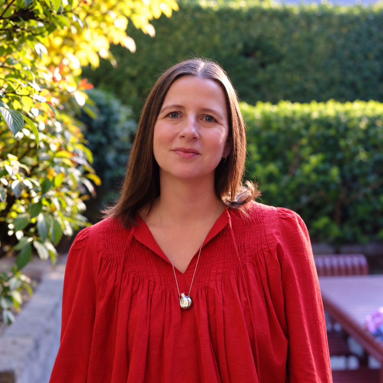 A woman with shoulder-length brown hair wearing a red blouse with pleated details and a silver pendant necklace, standing outdoors in a garden with green foliage and sunlight.