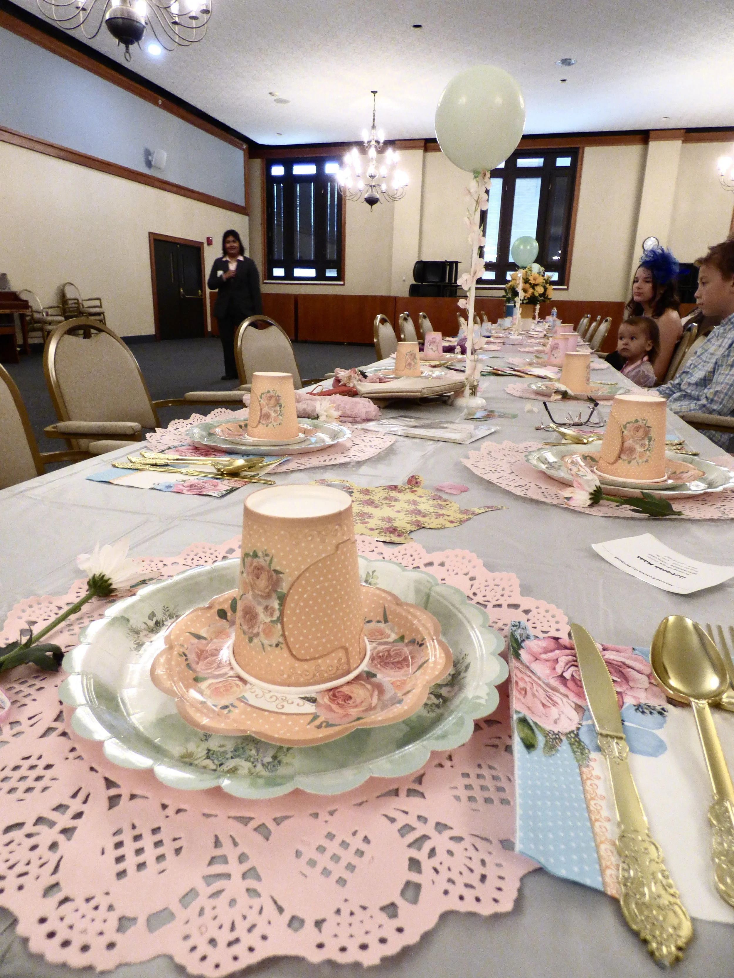 Decorated party table with pink paper doilies, floral paper cups with pink hearts, gold utensils, pink paper napkins, and floral-themed paper plates. A large balloon with pink and white flowers is attached to the center of the table. In the background, people are seated at the table, and a woman stands near the back wall of the event room.