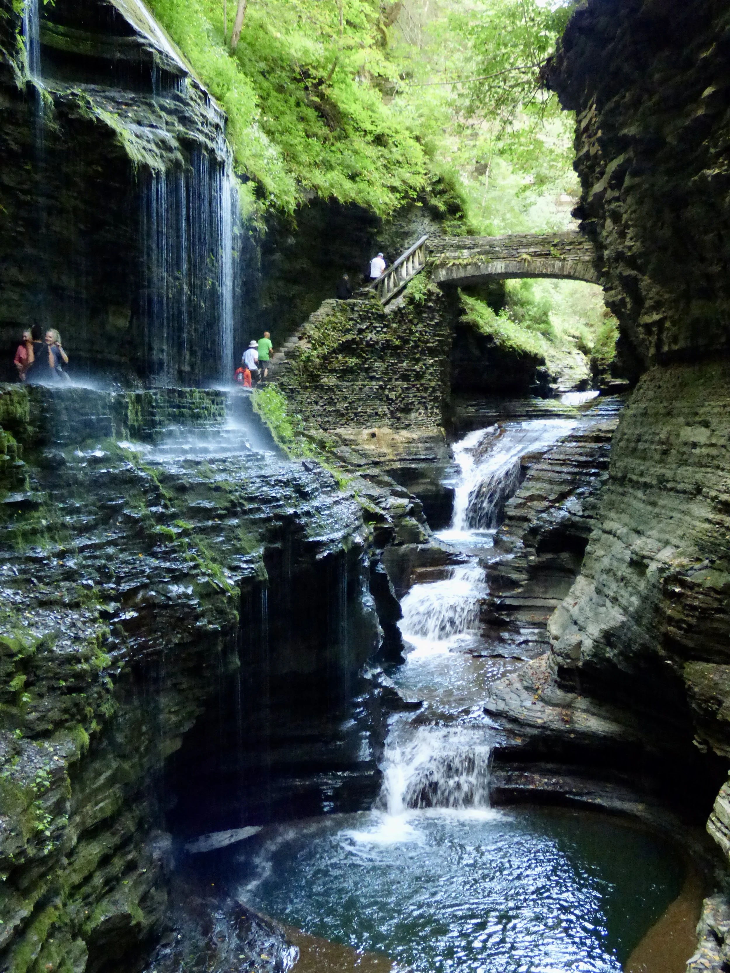 A narrow canyon with a small waterfall on the left side, greenery overhead, a stone bridge in the middle, and a stream flowing through the rocks at the bottom. Several people are hiking along a stone staircase and path near the waterfall.