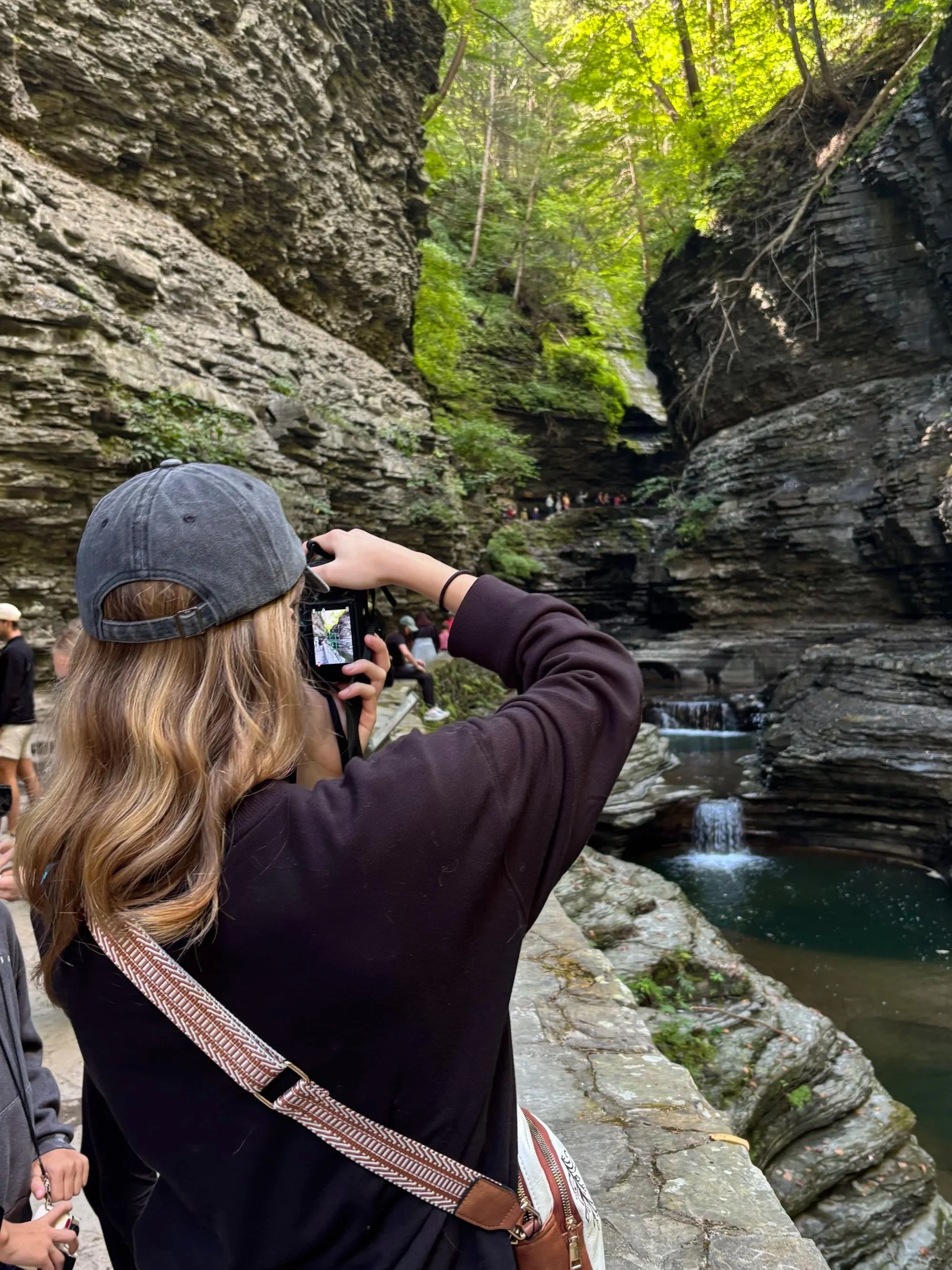 A person with long wavy blonde hair, wearing a gray cap and a dark jacket, is taking a photograph of a canyon with a small waterfall and lush green trees overhead. There are other people in the background near the canyon.