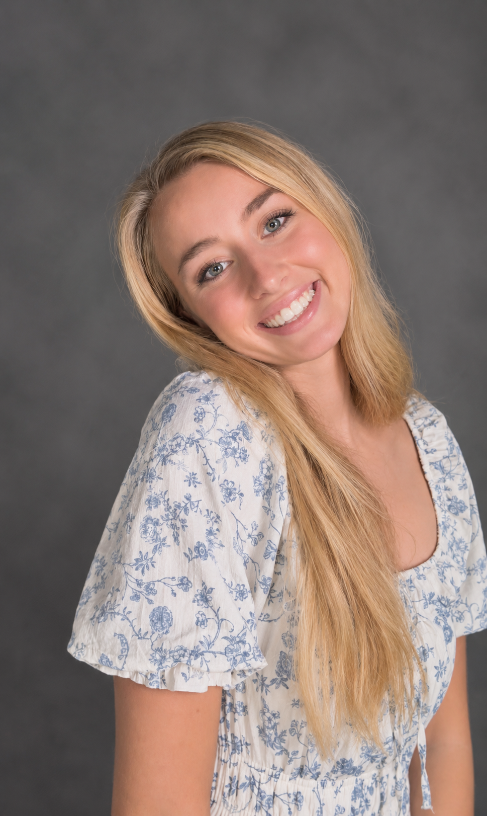 A young woman with blonde hair and blue eyes smiling, wearing a white dress with blue floral patterns against a dark gray background.