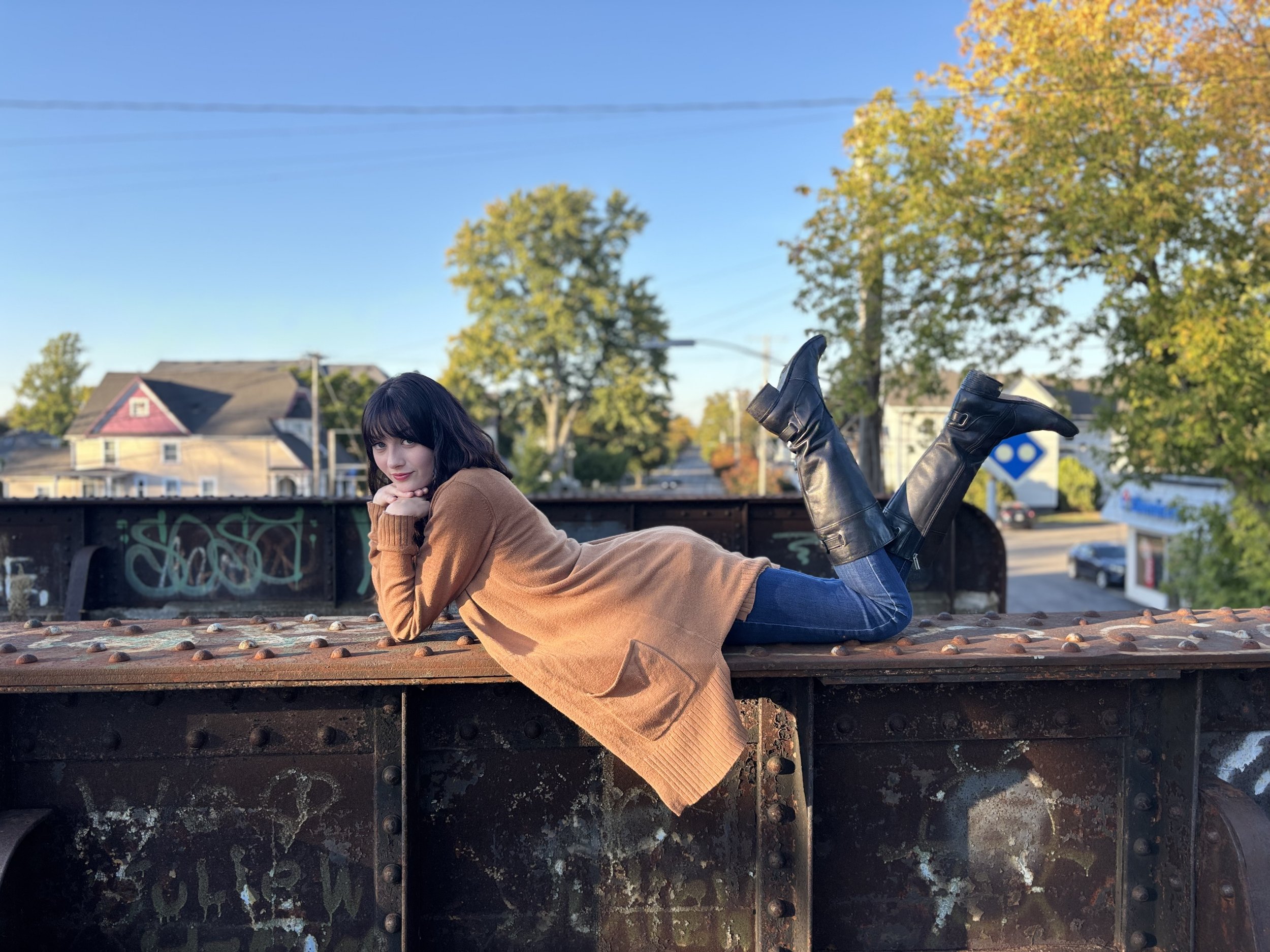 A woman with dark hair and bangs lying on her stomach on a metal bridge, wearing a brown coat and black leather boots, with trees and houses in the background on a clear day.