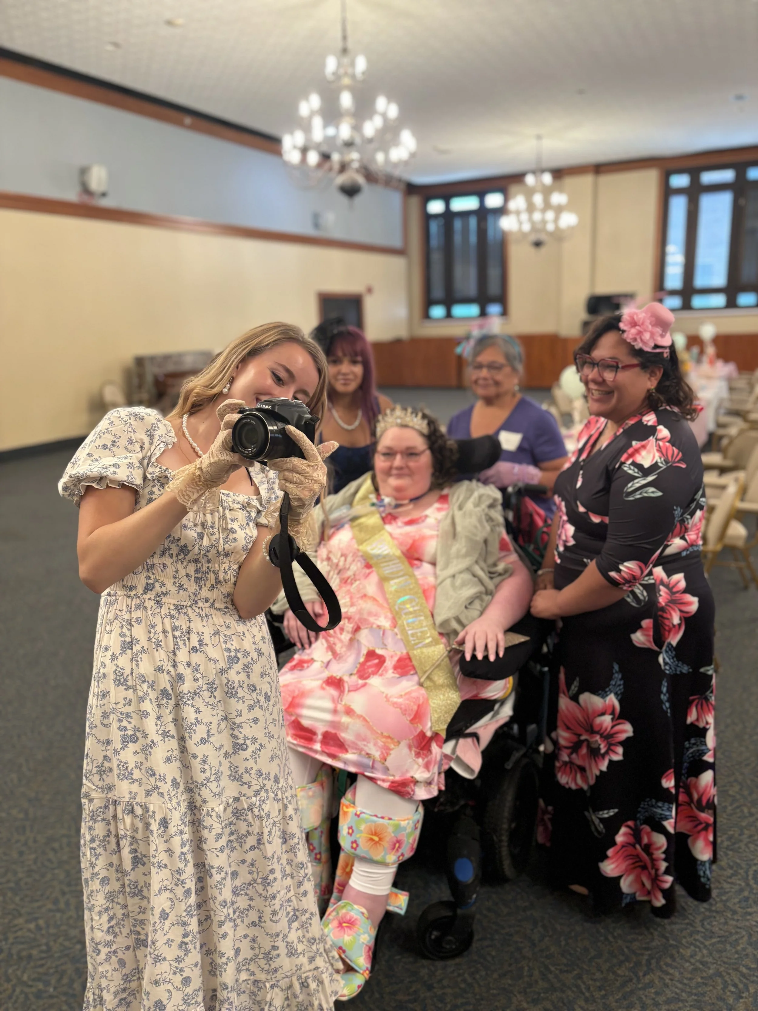 A group of women gathered in a room with chandeliers and large windows. One woman in a floral dress is taking a photo with a camera, while others in the group are smiling and dressed in colorful, festive clothing. One woman is wearing a sash and a floral headpiece, indicating a celebration.