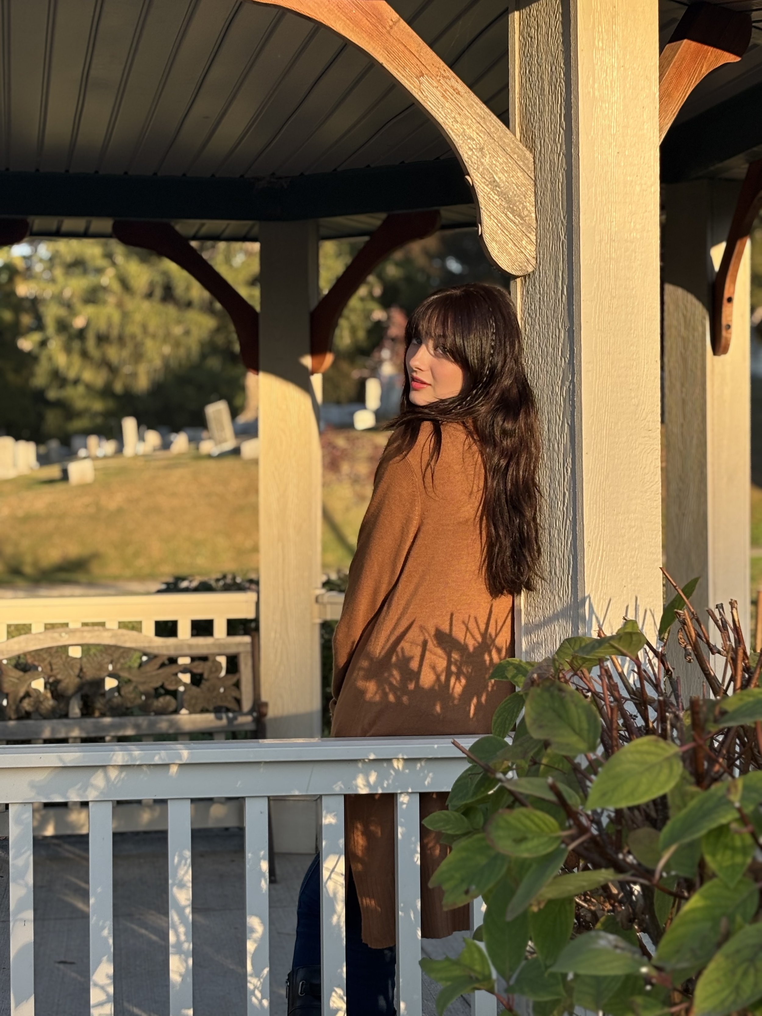 A woman with dark brown hair and a brown coat leaning against a wooden post on a porch, with sunlight casting shadows and green foliage in the background.