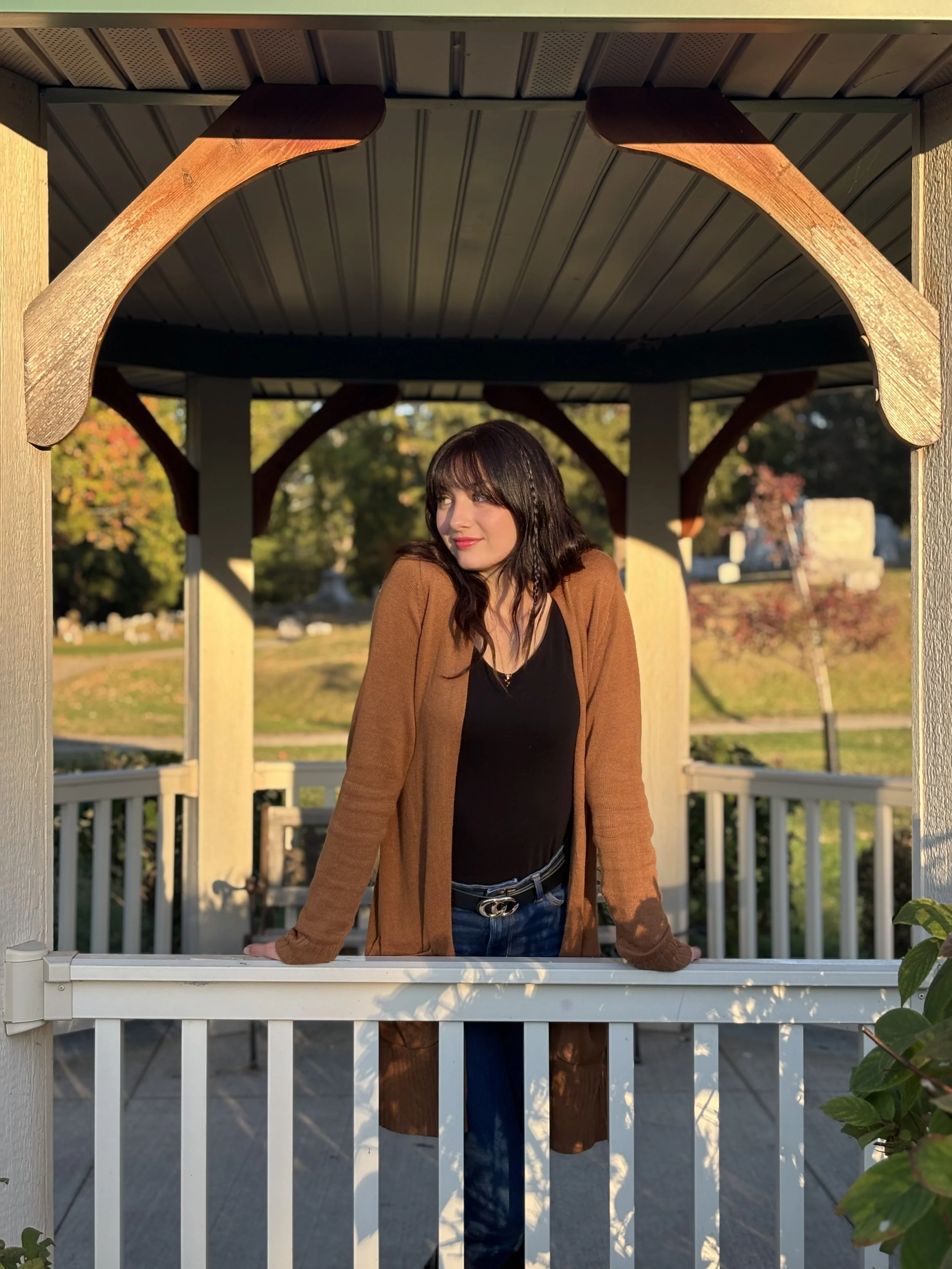 A woman with dark hair and a brown cardigan leaning on a white fence, standing under a gazebo with wooden supports, with trees and a park in the background during what appears to be late afternoon or early evening.
