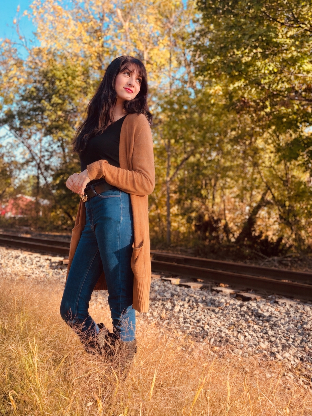 A woman with dark hair and fair skin stands outdoors near train tracks, surrounded by fall foliage with yellow and green leaves, wearing a long brown cardigan, black top, blue jeans, and boots, looking thoughtfully into the distance.