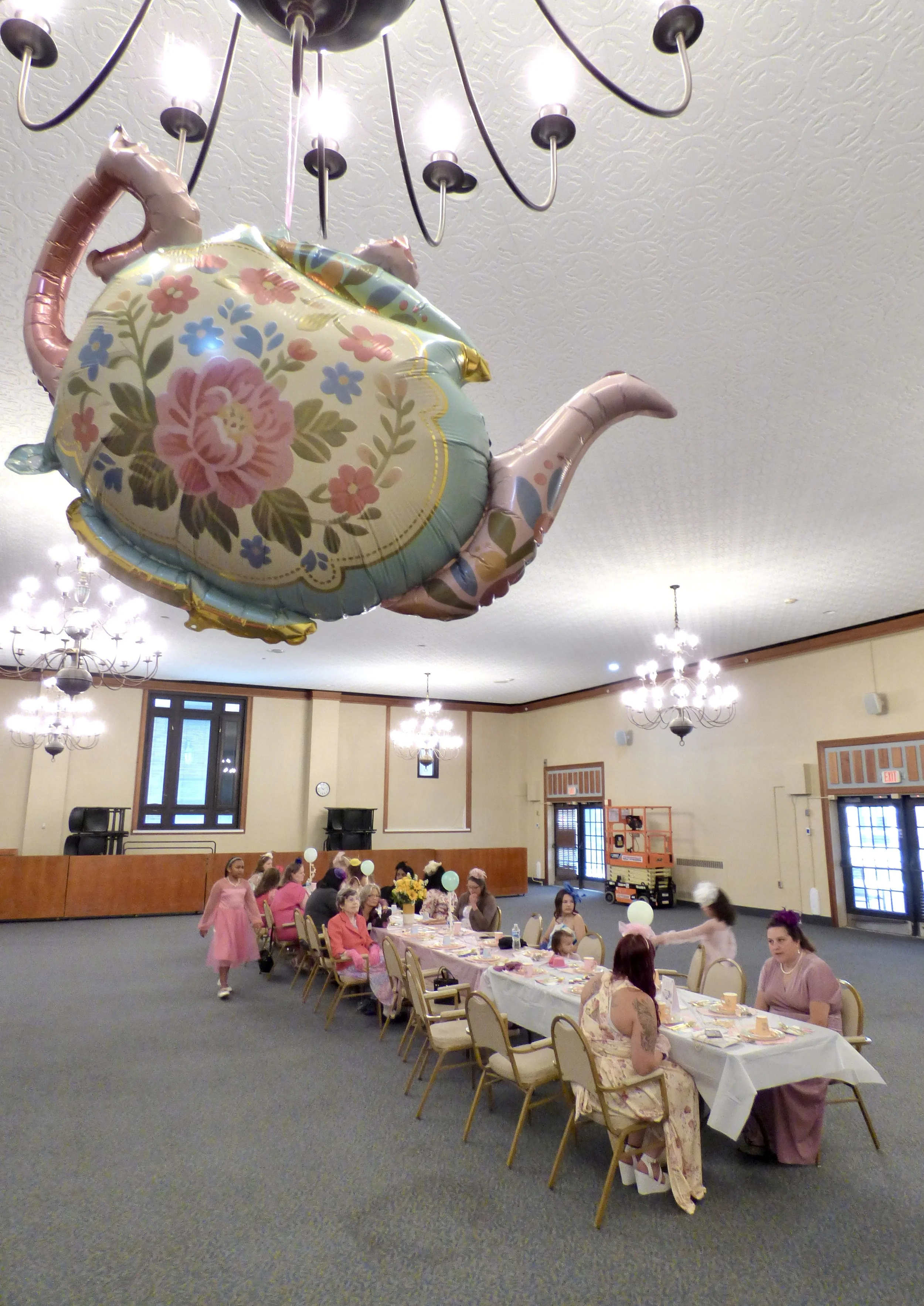 Children and adults gathered around a long, decorated dinner table at a party. A large pastel-colored teapot-shaped balloon with floral patterns floats above the table. The room has chandeliers, windows, and a process with a scissor lift in the background.