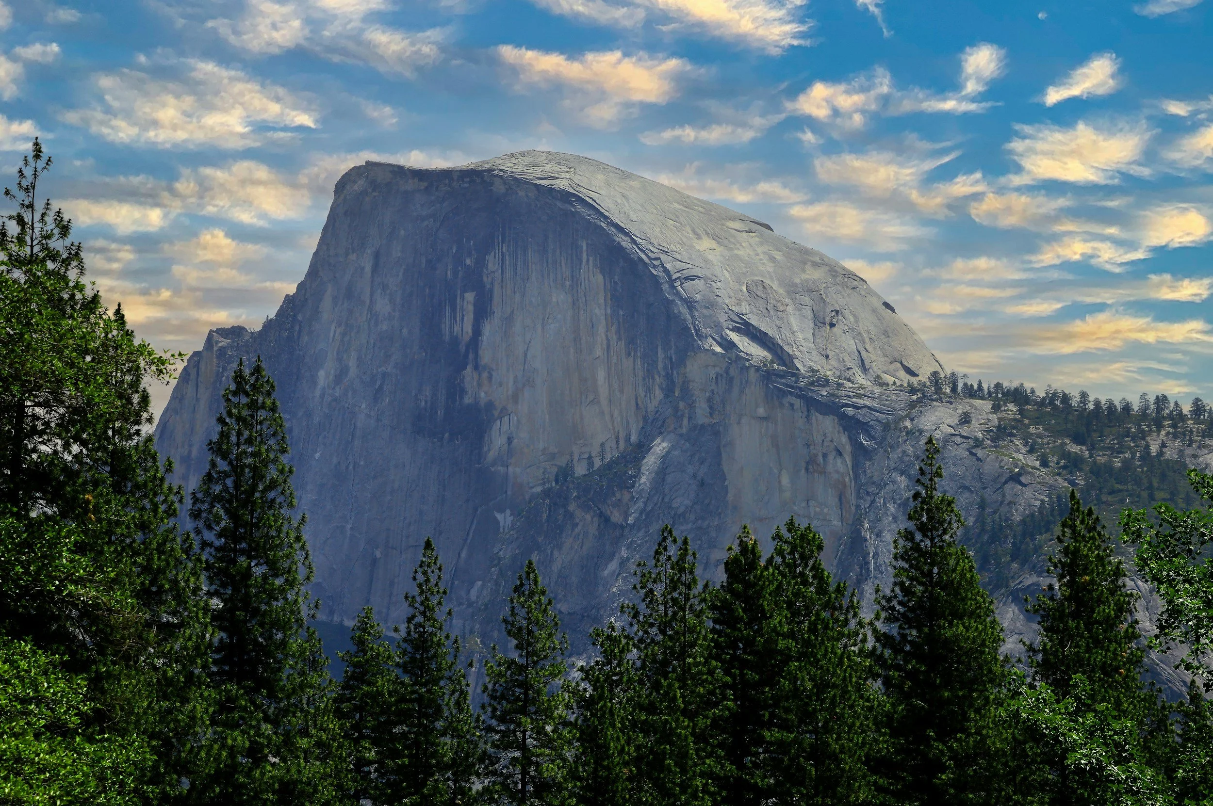 View of a large, granite mountain with a flat top, surrounded by evergreen trees, under a partly cloudy sky.