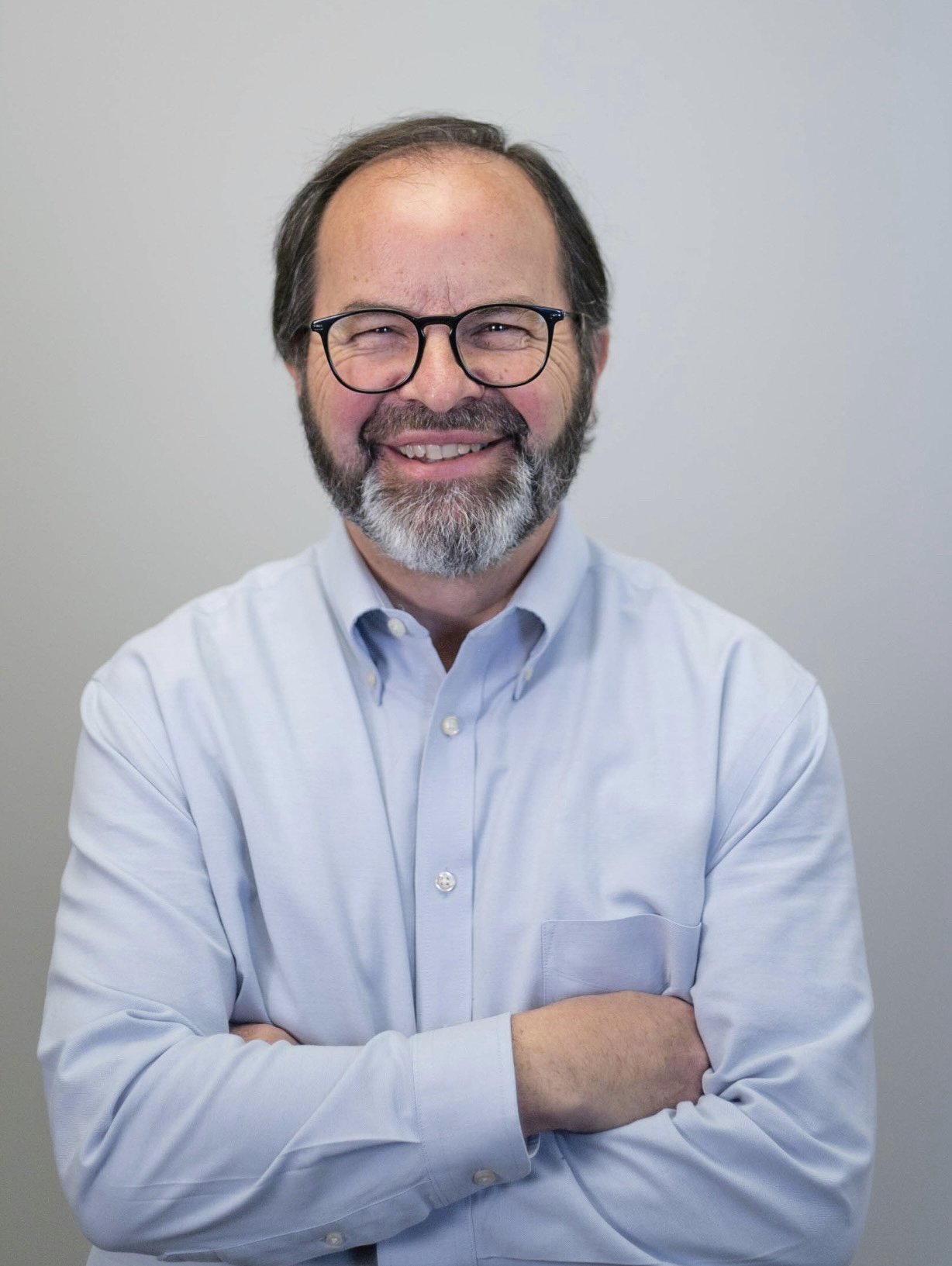 Man with glasses and beard, smiling, wearing a light blue button-up shirt with arms crossed, standing against a neutral background.