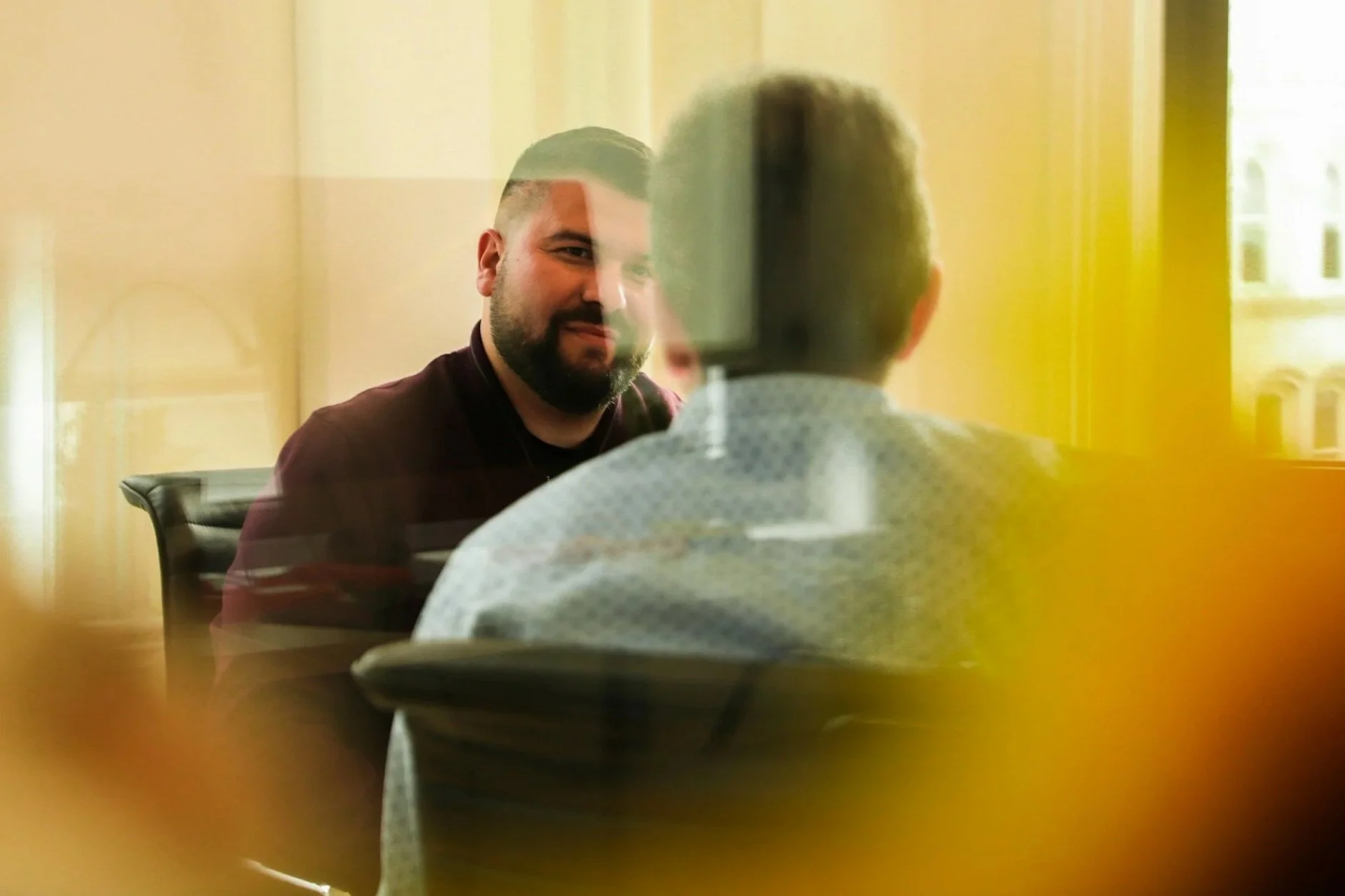 Two men having a conversation in an office, seen through a glass window with reflections and yellowish lighting.