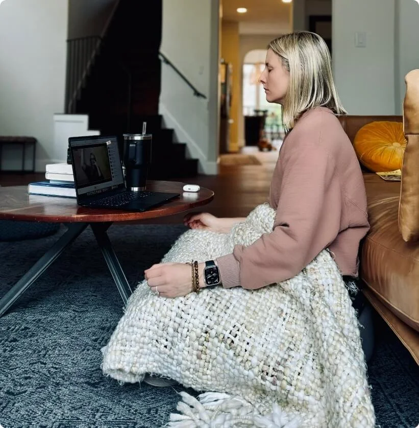 A woman sitting on the floor in a living room, facing a laptop on a coffee table, doing a guided breathwork session from Our Breath Collective