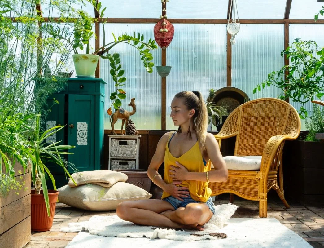A woman practicing breathwork indoors in a sunlit greenhouse or botanical space with plants, wicker chair, and decorative items.