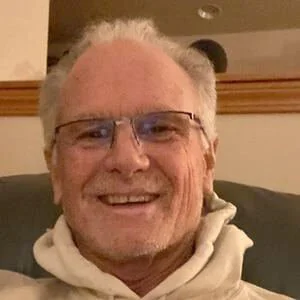 A smiling man with glasses and gray hair wearing a white hoodie, seated indoors.