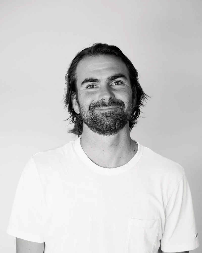 Black and white portrait of a man with medium-length dark hair, a full beard, wearing a plain white t-shirt, smiling softly, against a plain light gray background.