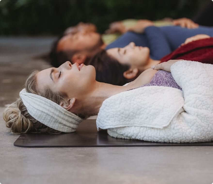Four people lying on their backs on yoga mats outdoors, eyes closed, practicing meditation or relaxation.