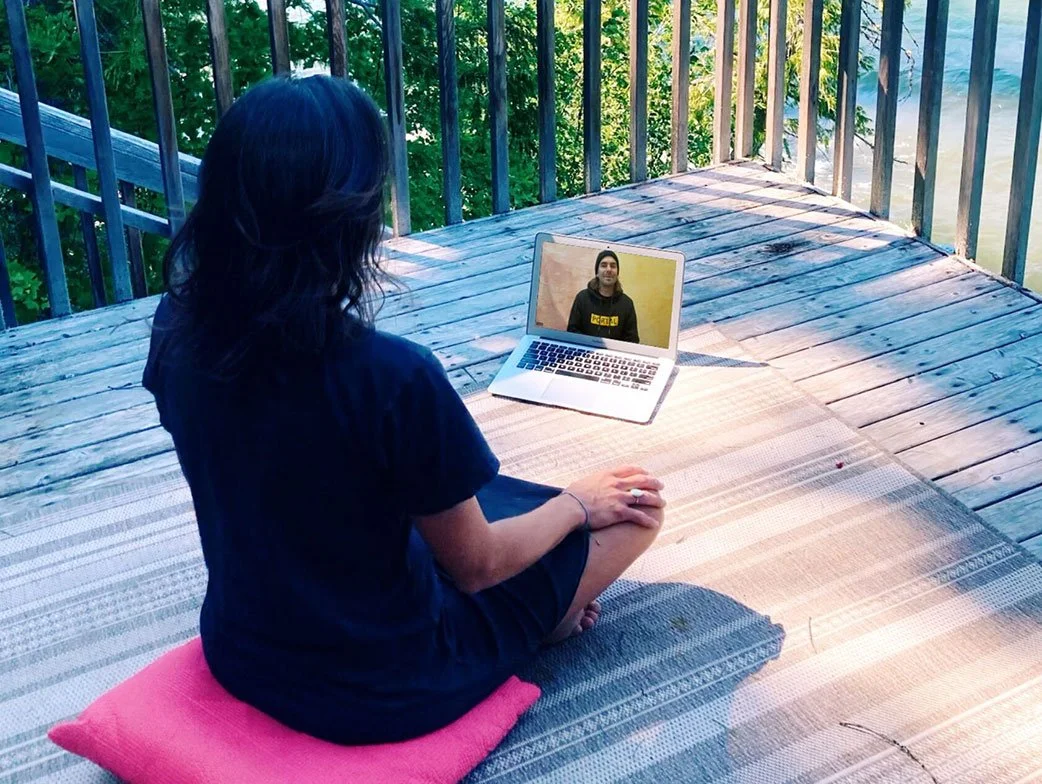 A woman with dark hair, dressed in dark clothing, sitting cross-legged on a pink cushion on a wooden deck, participating in a video call on her laptop. The deck is outdoors with a view of green trees and water.