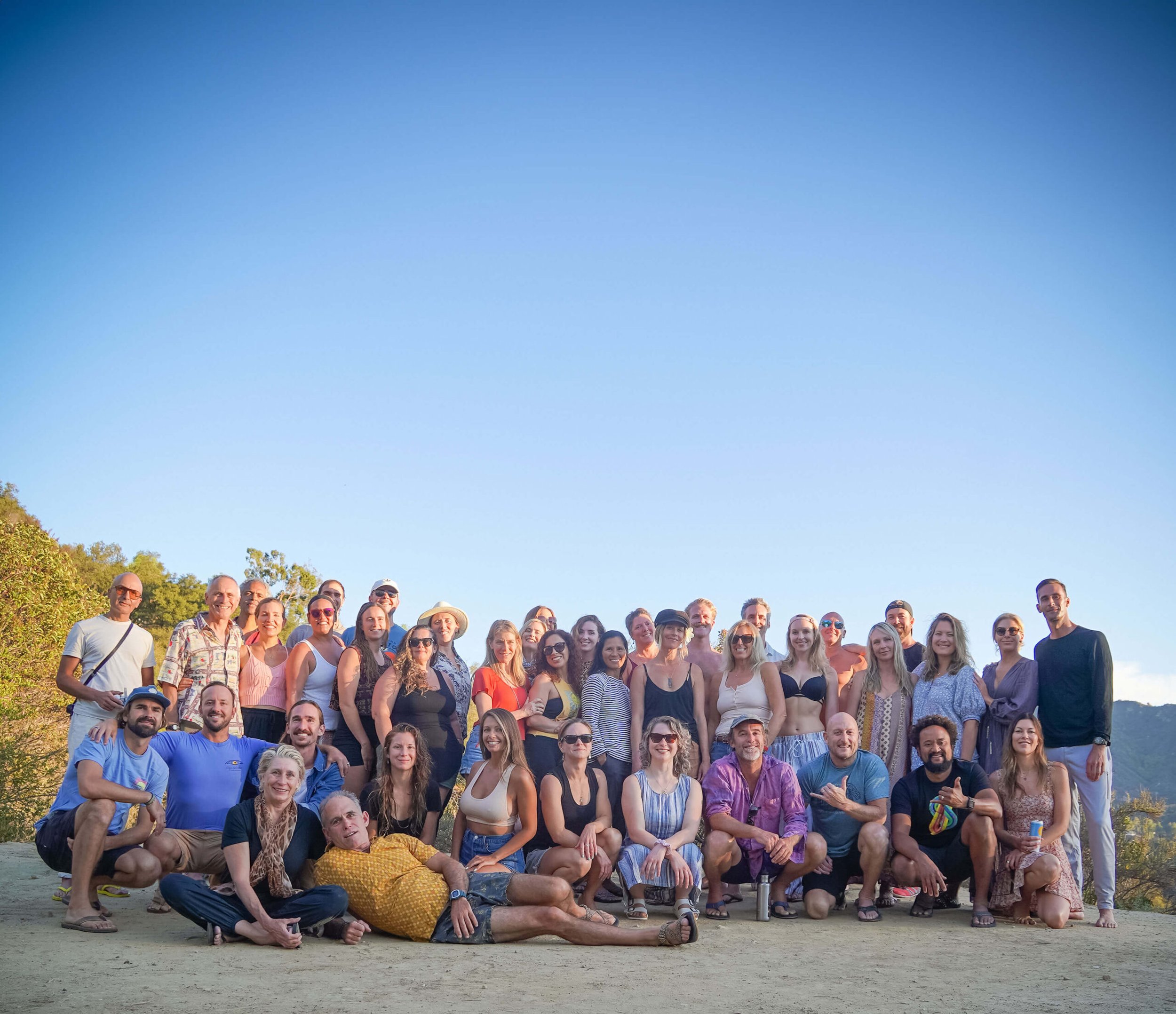 Large group of people posing outdoors on a sandy surface under a clear blue sky, with greenery and trees in the background.