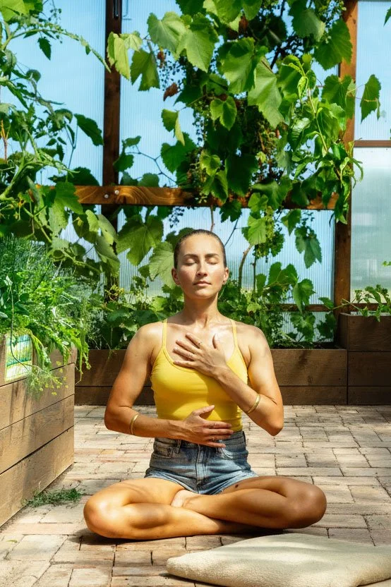 A woman practicing breathwork outdoors in a greenhouse surrounded by green plants, sitting cross-legged with eyes closed and hands placed on her chest and stomach.