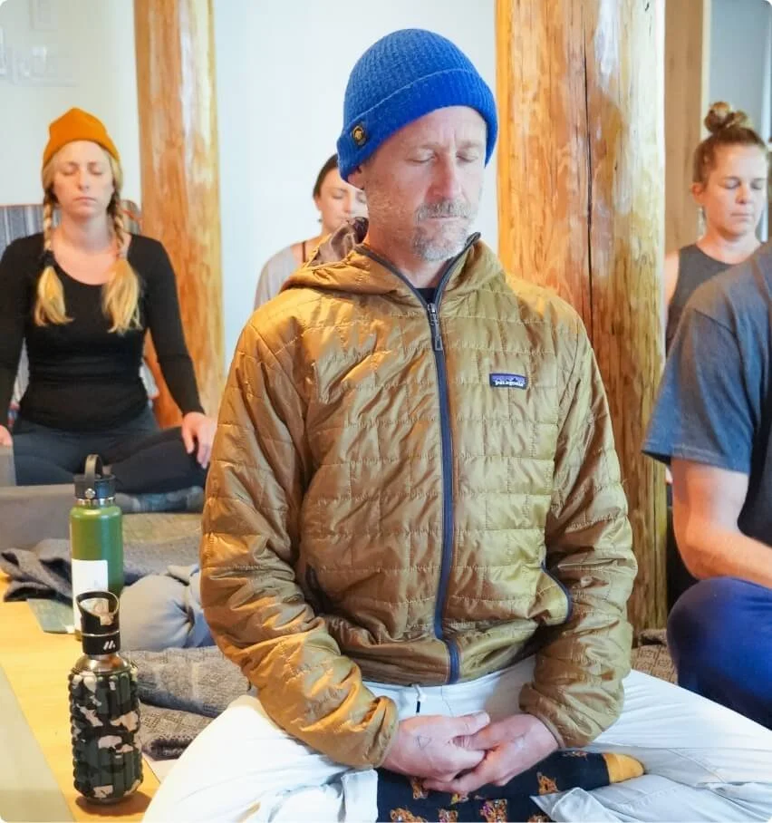 A man in a blue beanie and tan jacket sitting cross-legged with eyes closed and hands resting in his lap, meditating among others who are also meditating in a cozy, wood-paneled room.