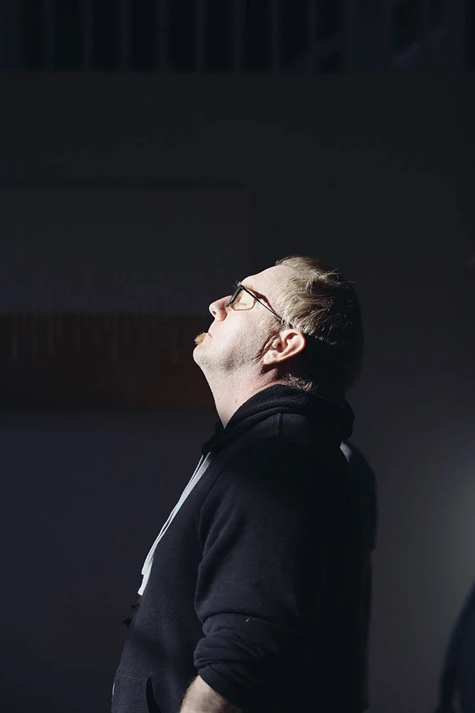 A man with glasses and a beard, wearing a black hoodie, stands with his head tilted back and eyes closed, possibly enjoying a moment of relaxation or reflection.