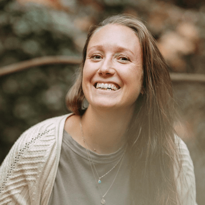 Smiling young woman outdoors with brown hair, wearing a gray shirt and beige cardigan, in a natural setting.