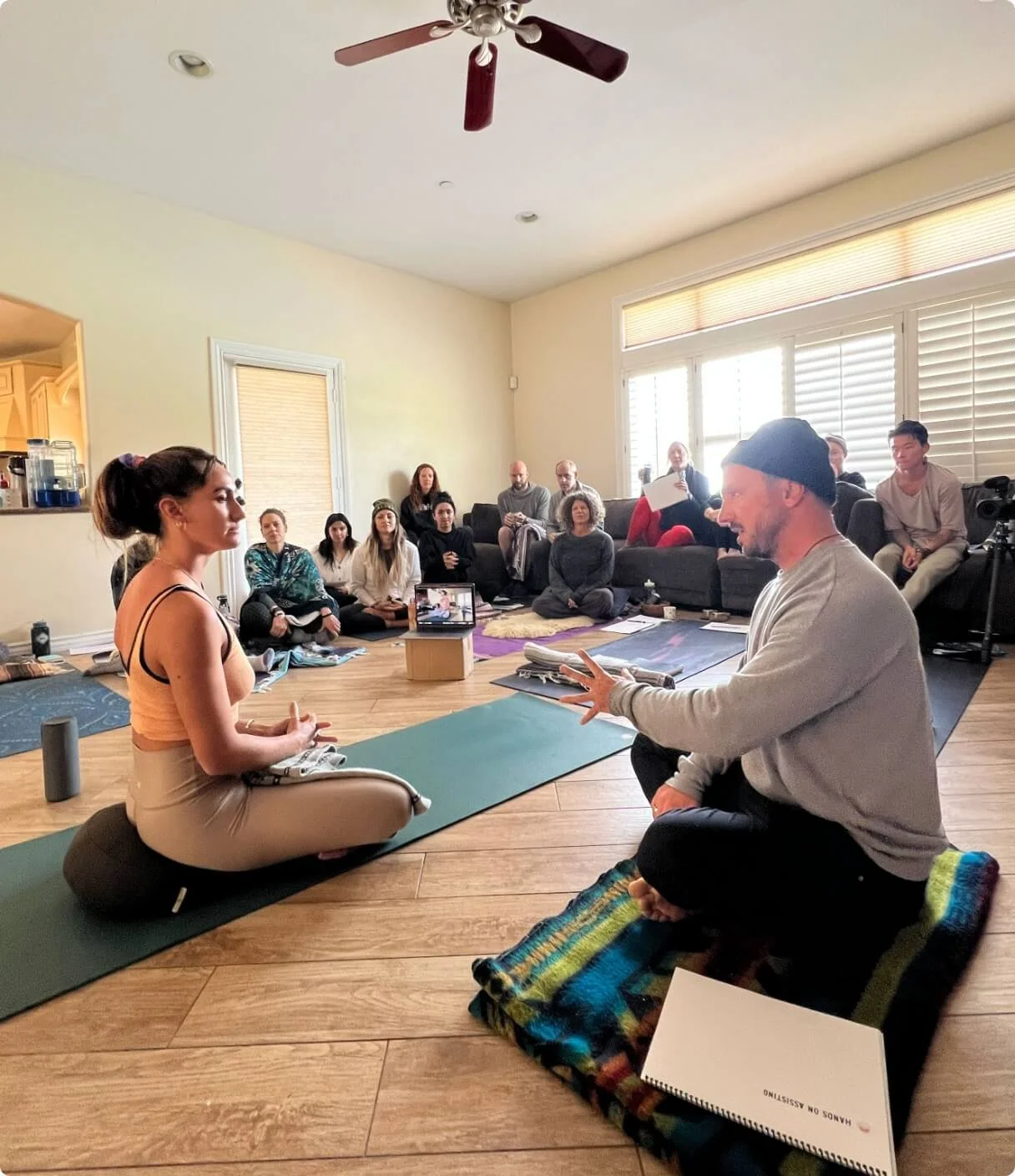 A group of people attending a meditation or yoga class in a spacious, well-lit room with wooden flooring. Two individuals, a woman and a man, are seated on yoga mats at the front, engaging in instruction or conversation. The woman is in a cross-legged meditative pose, while the man, wearing a beanie, appears to be explaining or leading. Several others sit on the floor or couches, observing or listening.