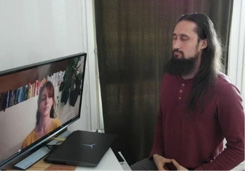 A man sitting with his eyes closed in front of a monitor displaying a woman leading an online breathwork session.