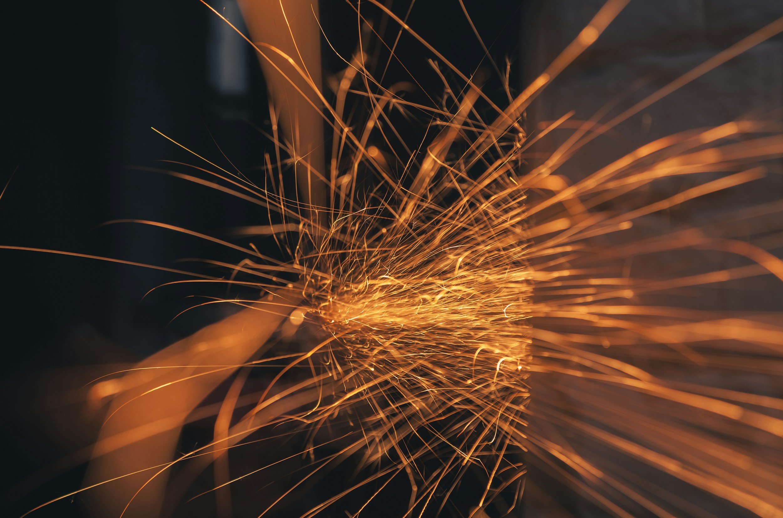 Close-up of sparks flying outward from a metallic surface as it is being ground or cut, with a dark background.