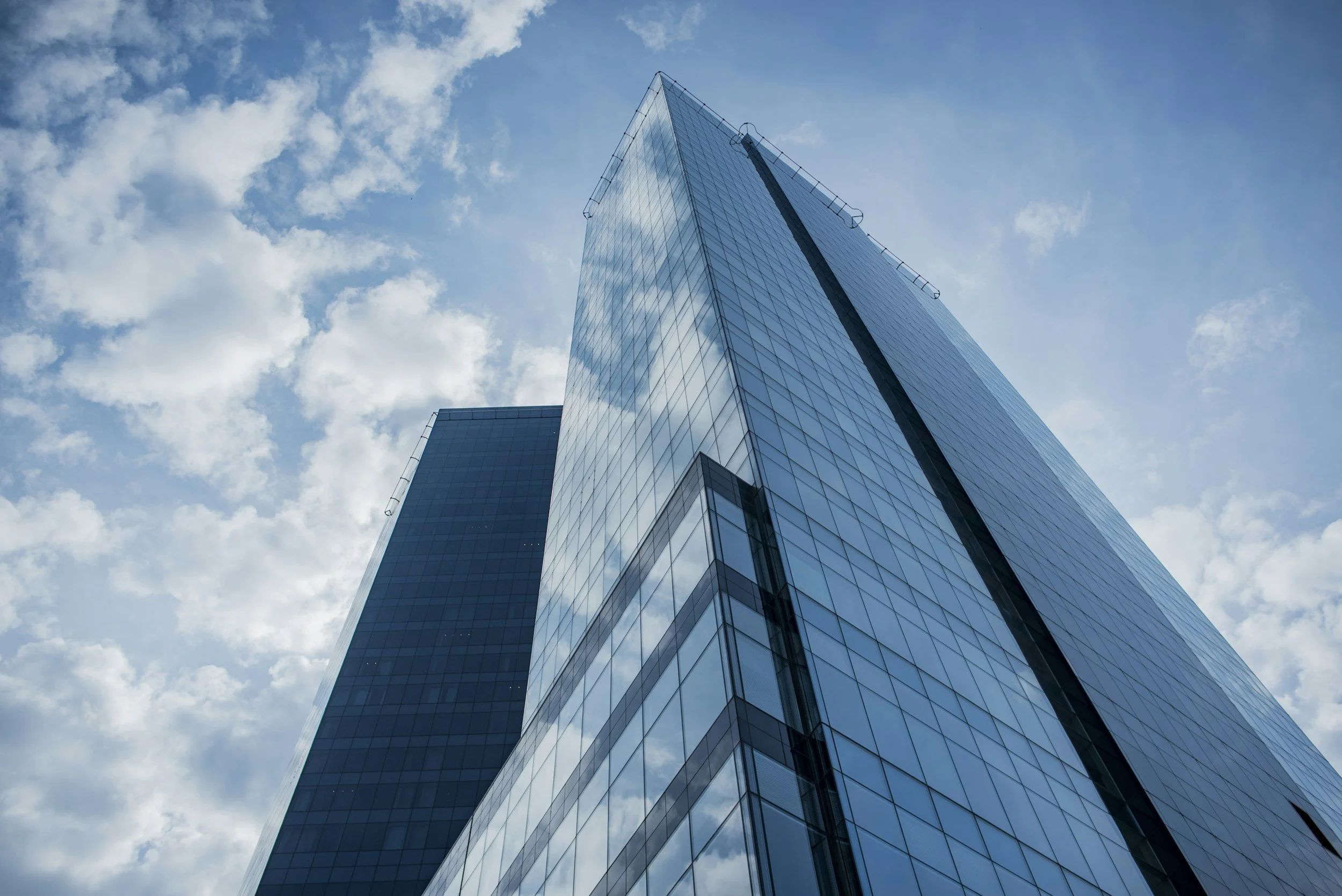 A tall modern glass skyscraper seen from below, with a partly cloudy blue sky in the background.