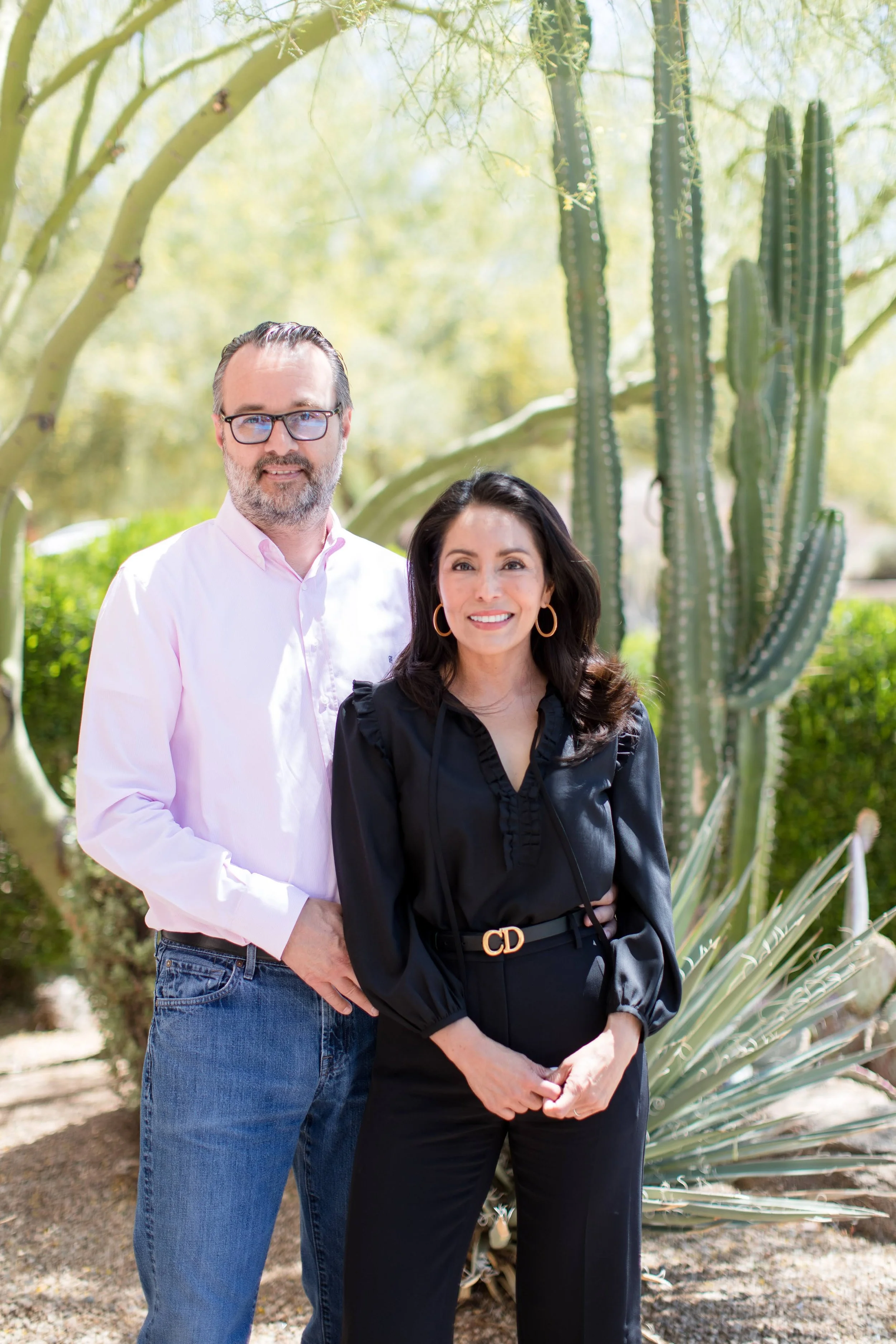A man with glasses and a beard wearing a light pink shirt and jeans stands next to a woman with dark hair wearing a black blouse and black pants, posing outdoors among cacti and desert plants.
