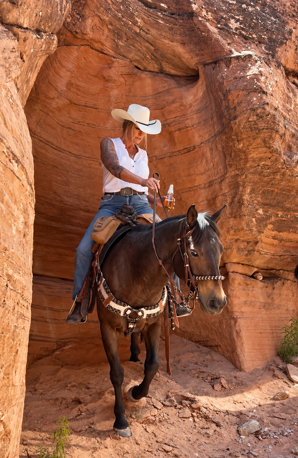 A woman wearing a white cowboy hat, white sleeveless shirt, and jeans is riding a horse through a narrow canyon with reddish rock walls. She holds a bottle and a stick in her hands.