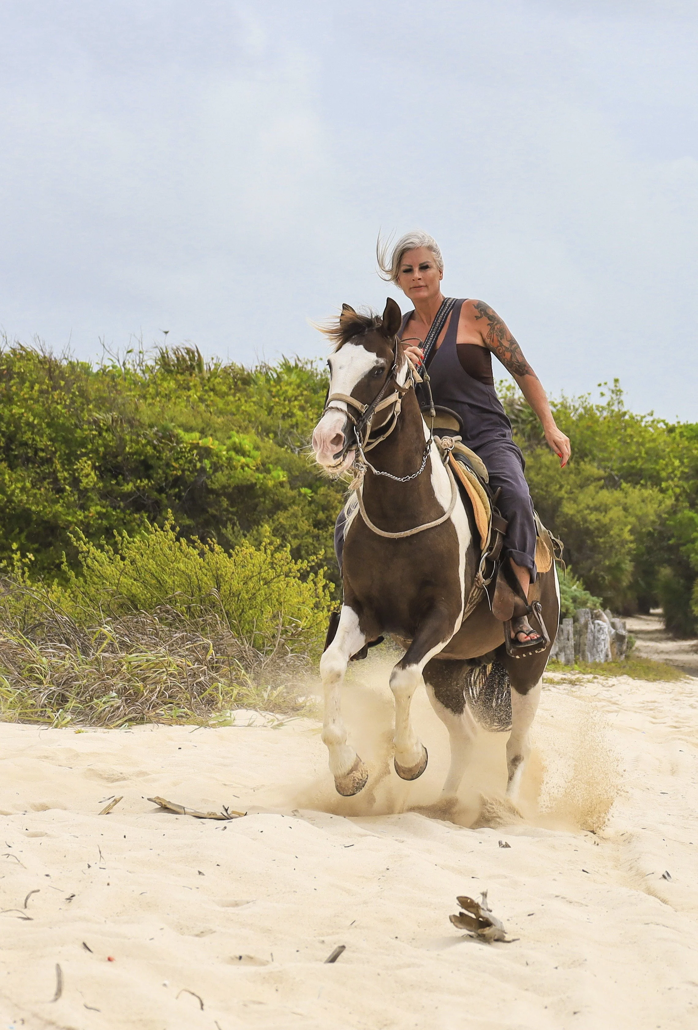 A woman riding a bucking horse on a sandy beach with green bushes and trees in the background.