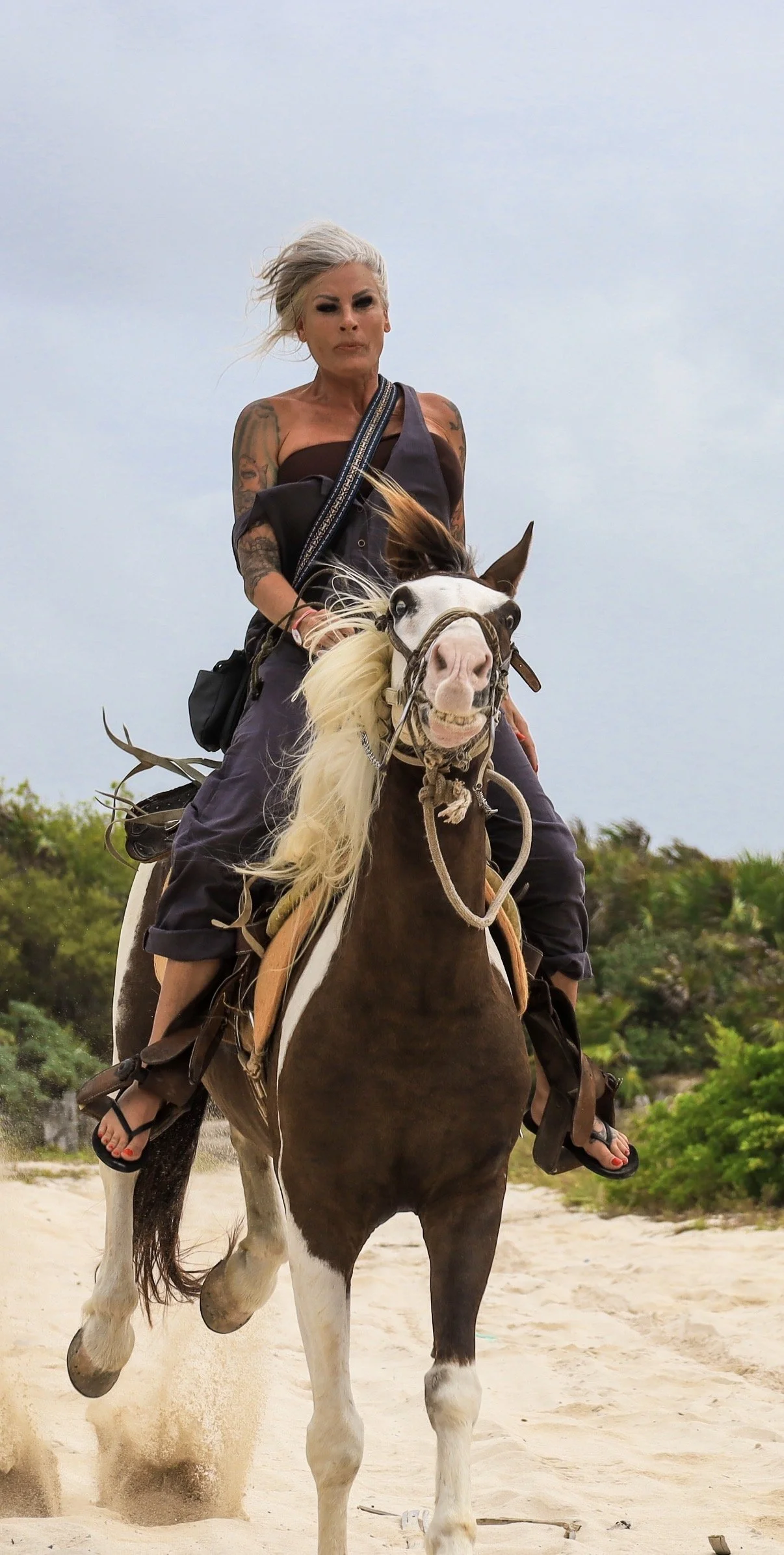 Woman with tattoos riding a pinto horse on the beach, blowing hair in the wind.