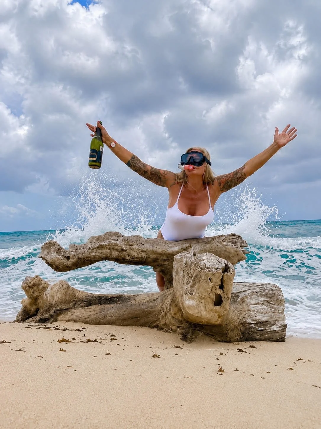 Woman in a white swimsuit with tattoos on her arms, wearing snorkeling gear and holding a champagne bottle, sitting on driftwood on the beach with ocean waves crashing behind her.