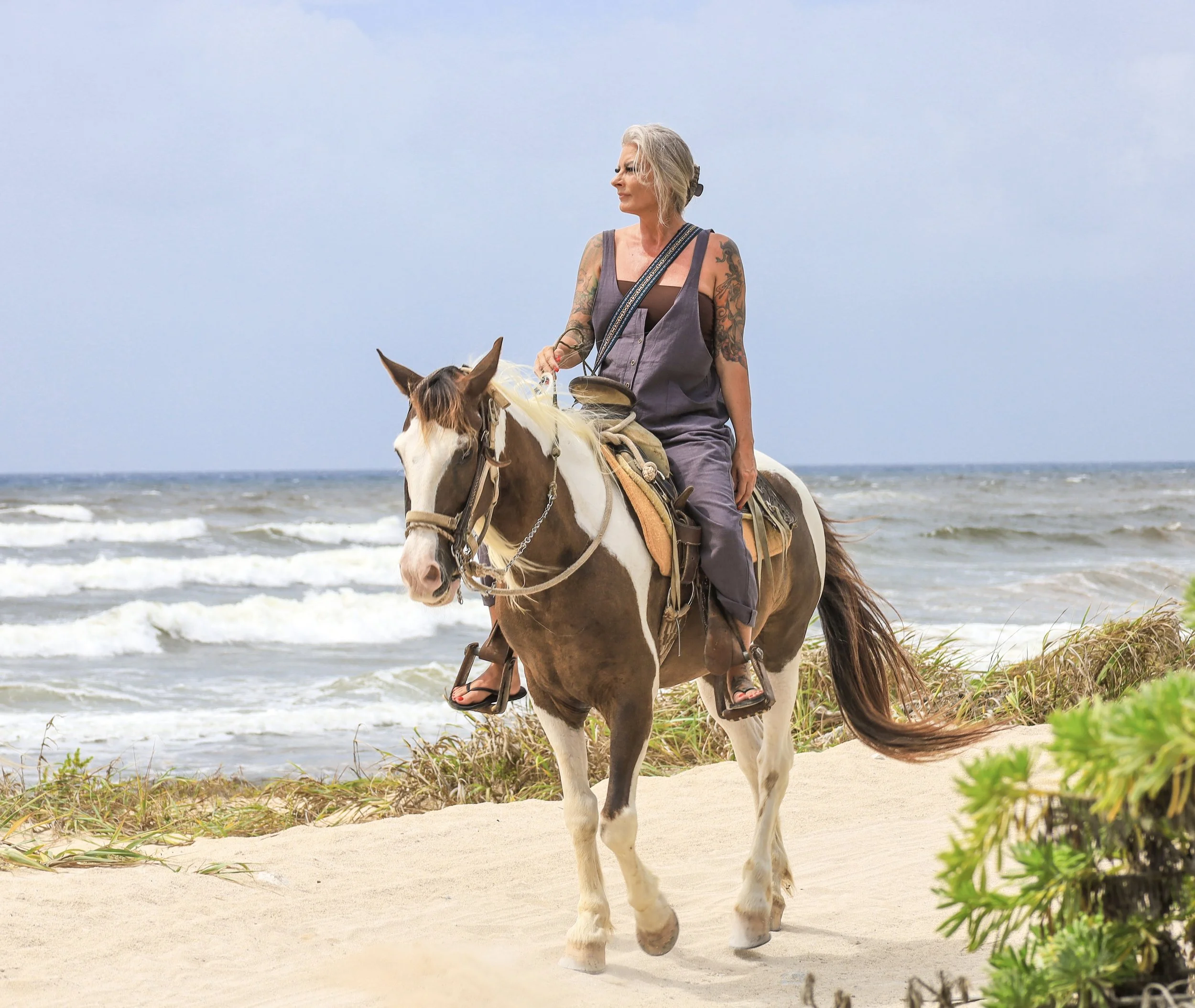 A woman with tattoos riding a horse along the beach, with the ocean waves and sandy shore in the background.