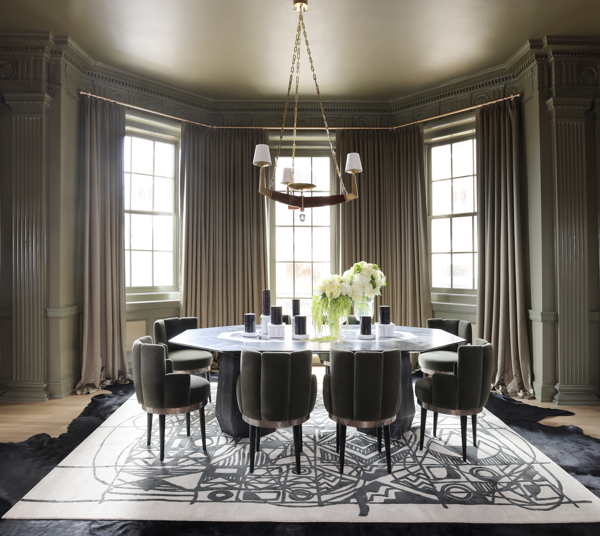 A dining room with large windows, surrounded by cream-colored walls with decorative molding, and curtains. A round table with a black base is set with multiple black and white candles and a large bouquet of white and green flowers. Six dark upholster