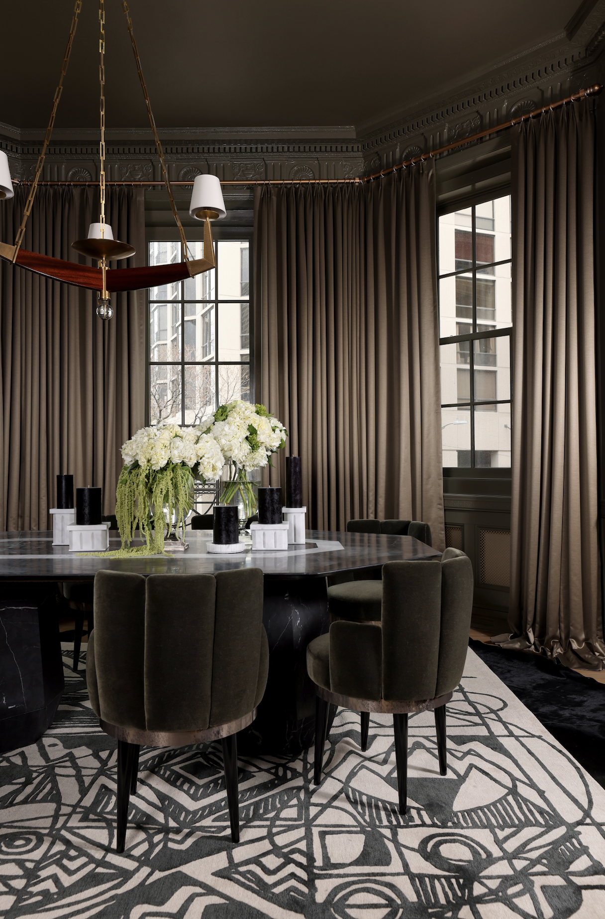 Elegant dining room with a round table, surrounded by green upholstered chairs, decorated with a floral centerpiece, large windows with beige curtains, and a geometric patterned rug.