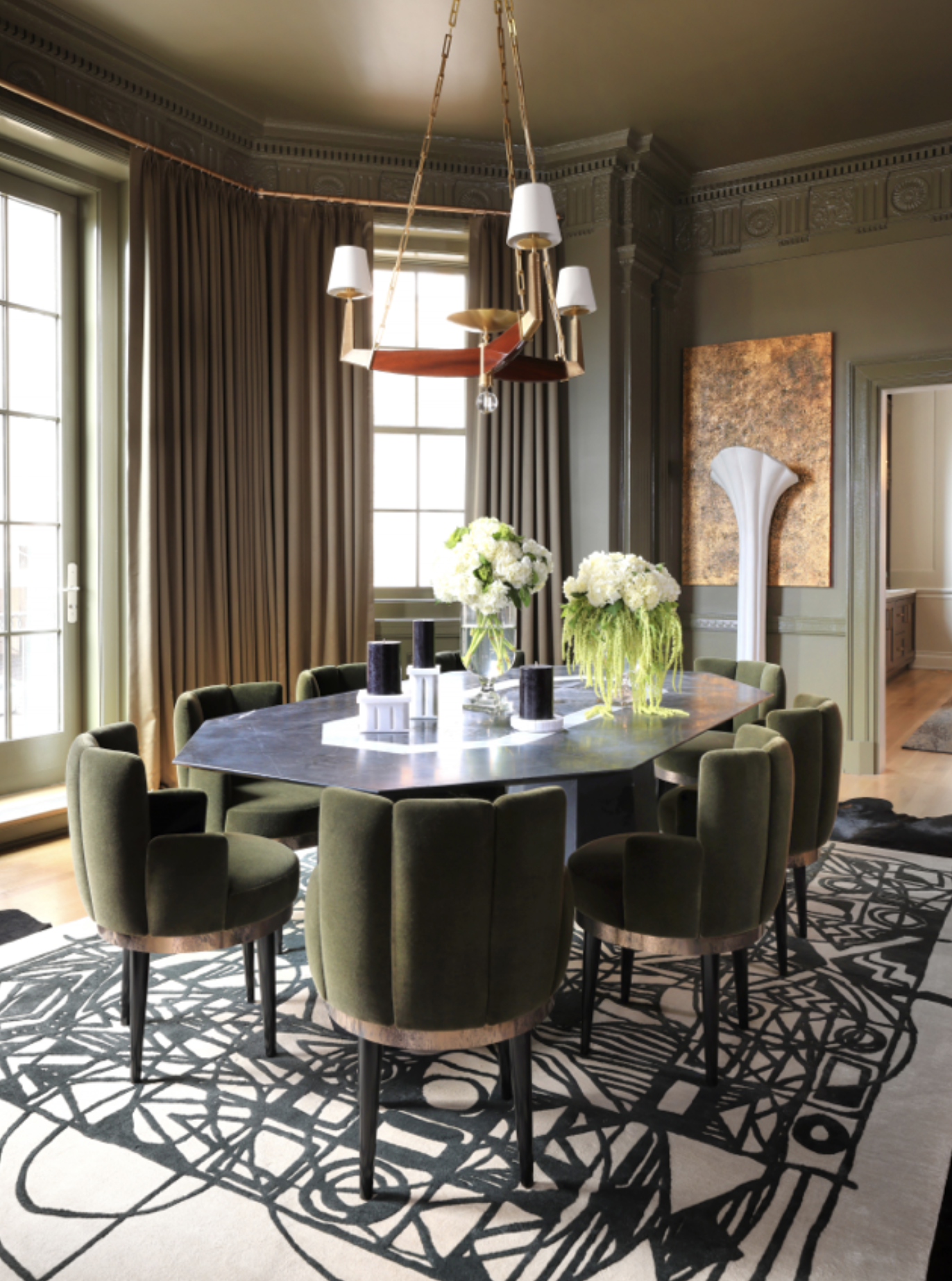 A dining room with a round table surrounded by green upholstered chairs, a black and white patterned rug, tall windows with gold curtains, and a modern chandelier hanging from the ceiling, with floral arrangements on the table.