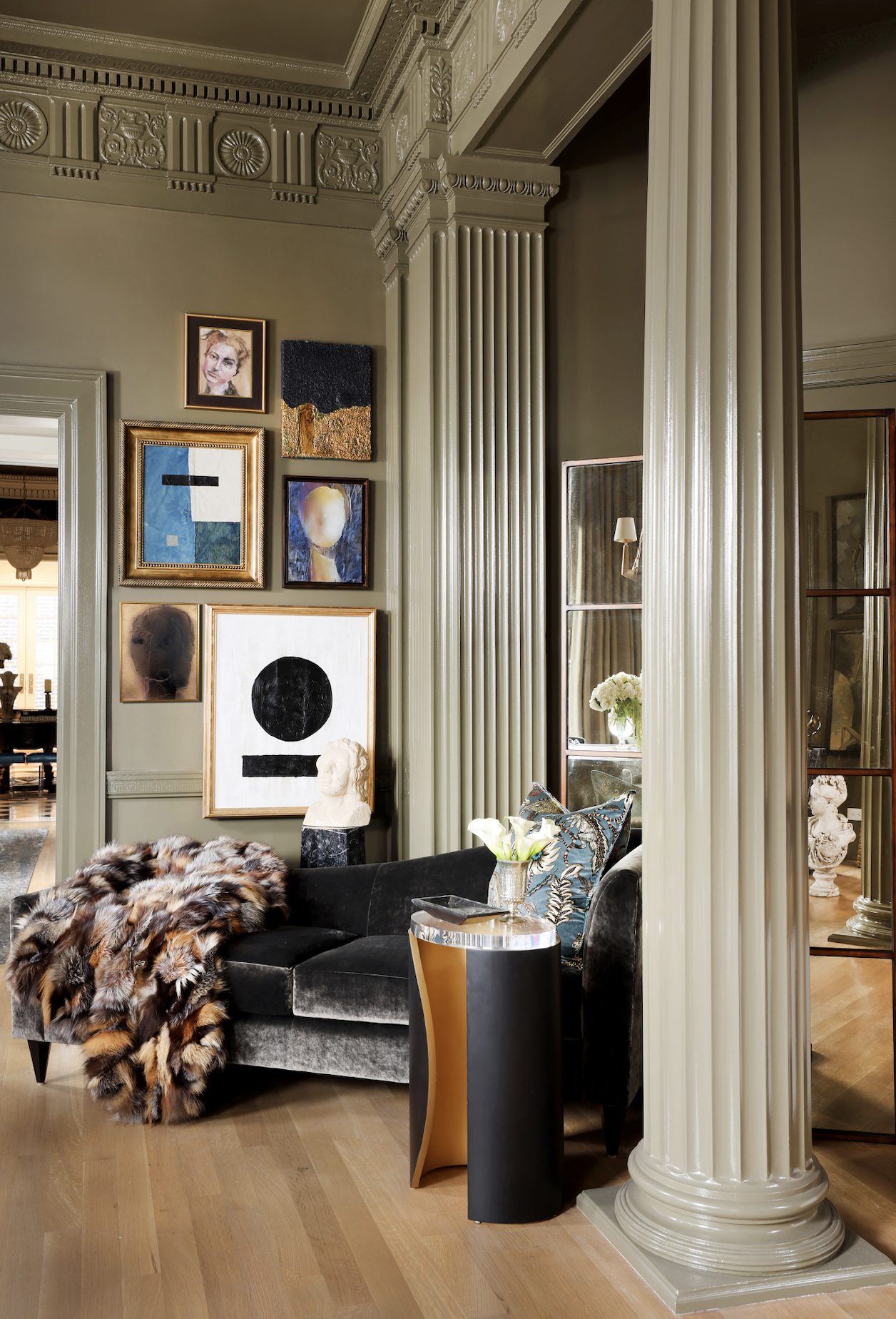Living room with ornate molding, contemporary artwork on the wall, a black velvet sofa with a fur throw, a small side table with a flower arrangement, and a classical column.