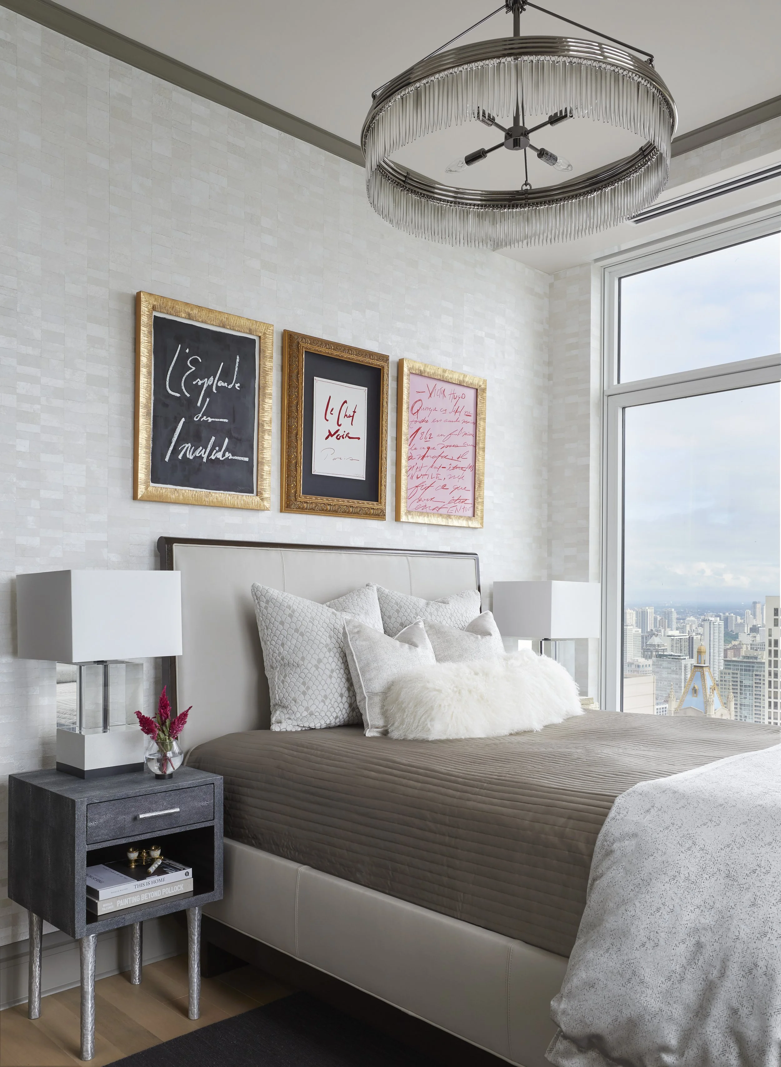 Modern bedroom with a large window view of a city skyline, decorated with framed artwork, a white headboard, and a side table with a small flower arrangement.