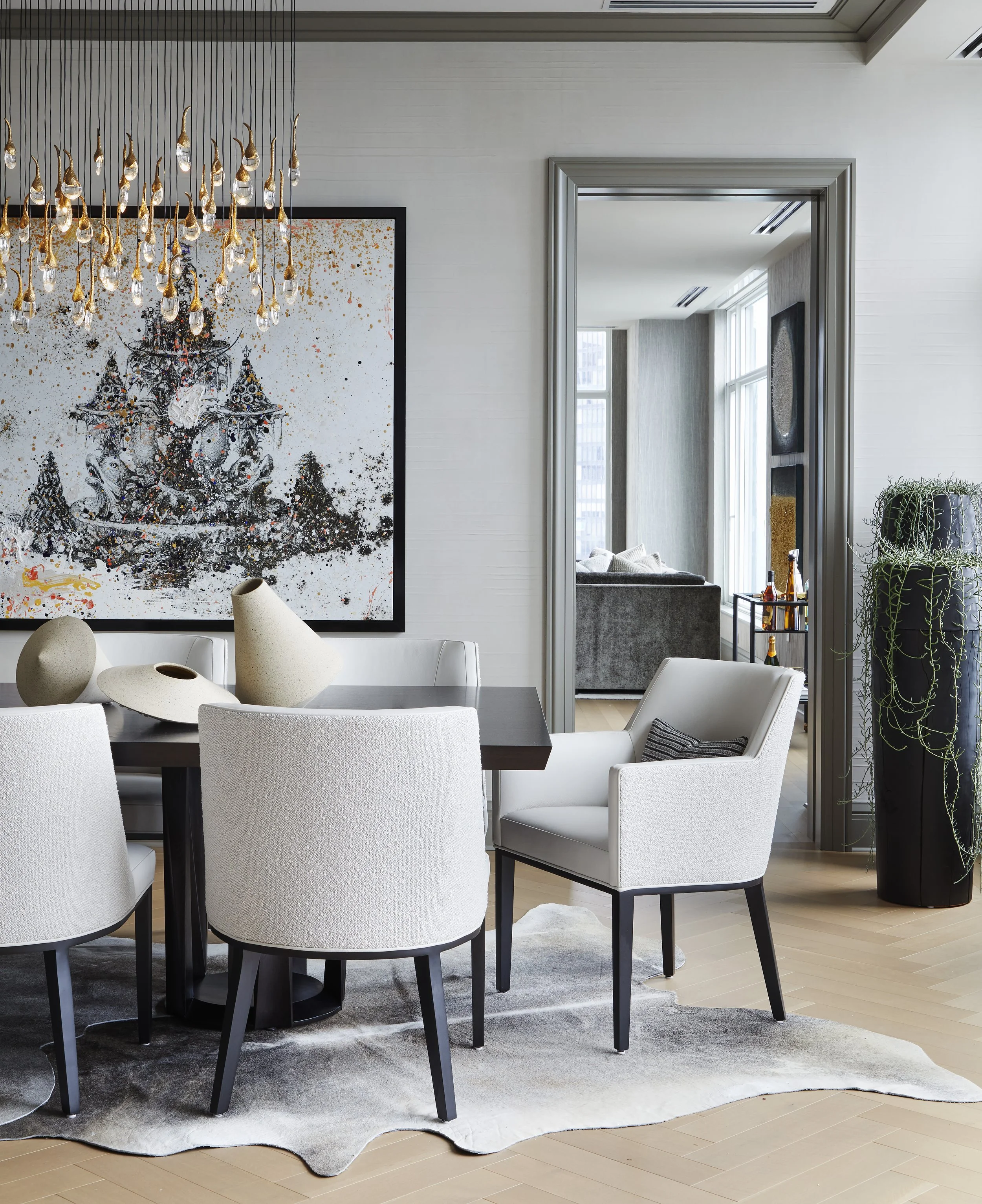 A modern dining area with a long black table surrounded by white upholstered chairs, positioned on a light hardwood floor with a gray rug underneath. There are decorative vases on the table, and the space features a large, colorful abstract artwork o