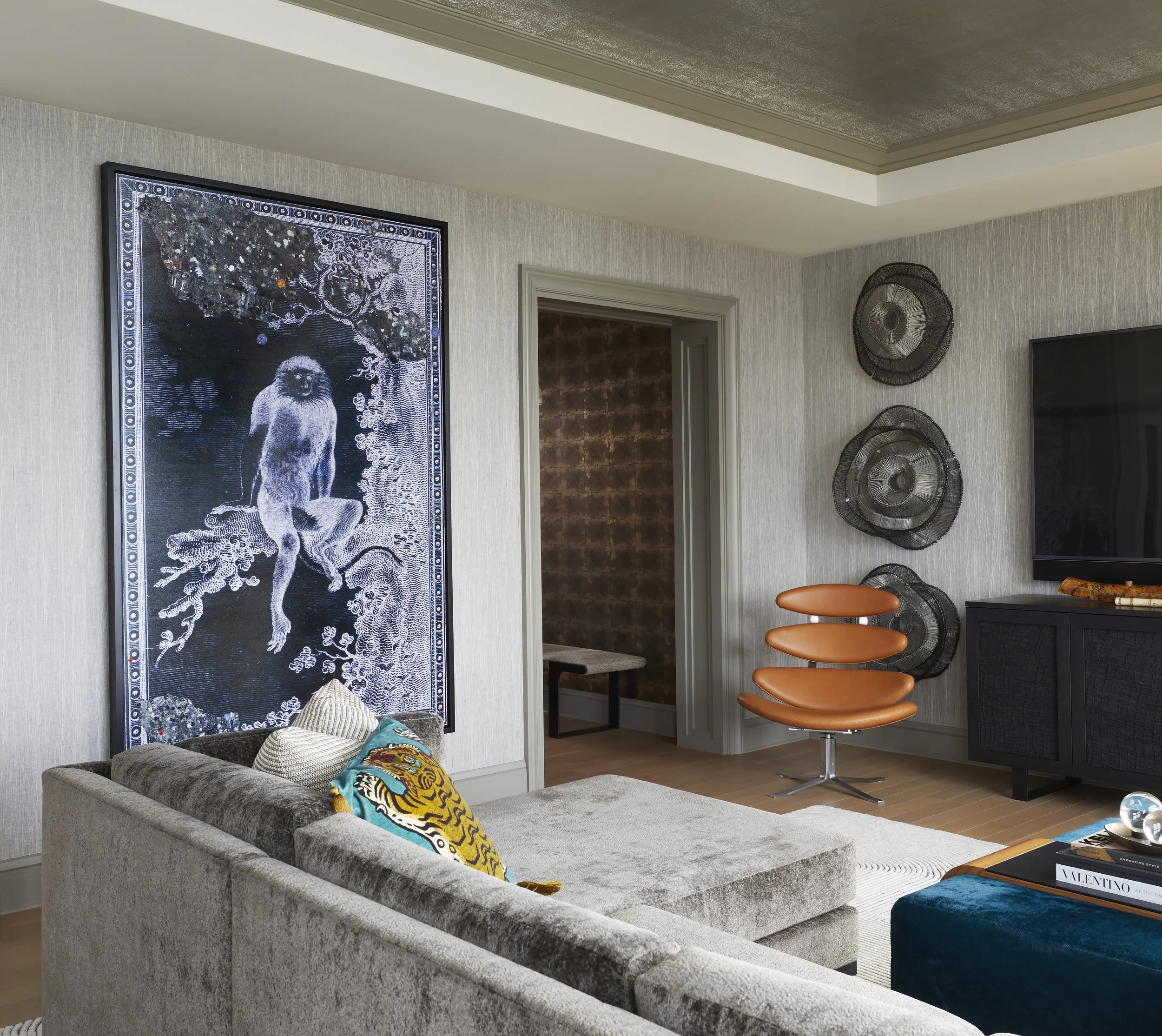 A living room corner with a gray velvet sofa, colorful throw pillows, a wall decorated with black wire circular wall art, a framed artwork featuring a monkey and a tree, a doorway leading to another room, and a partial view of a flat screen TV on the