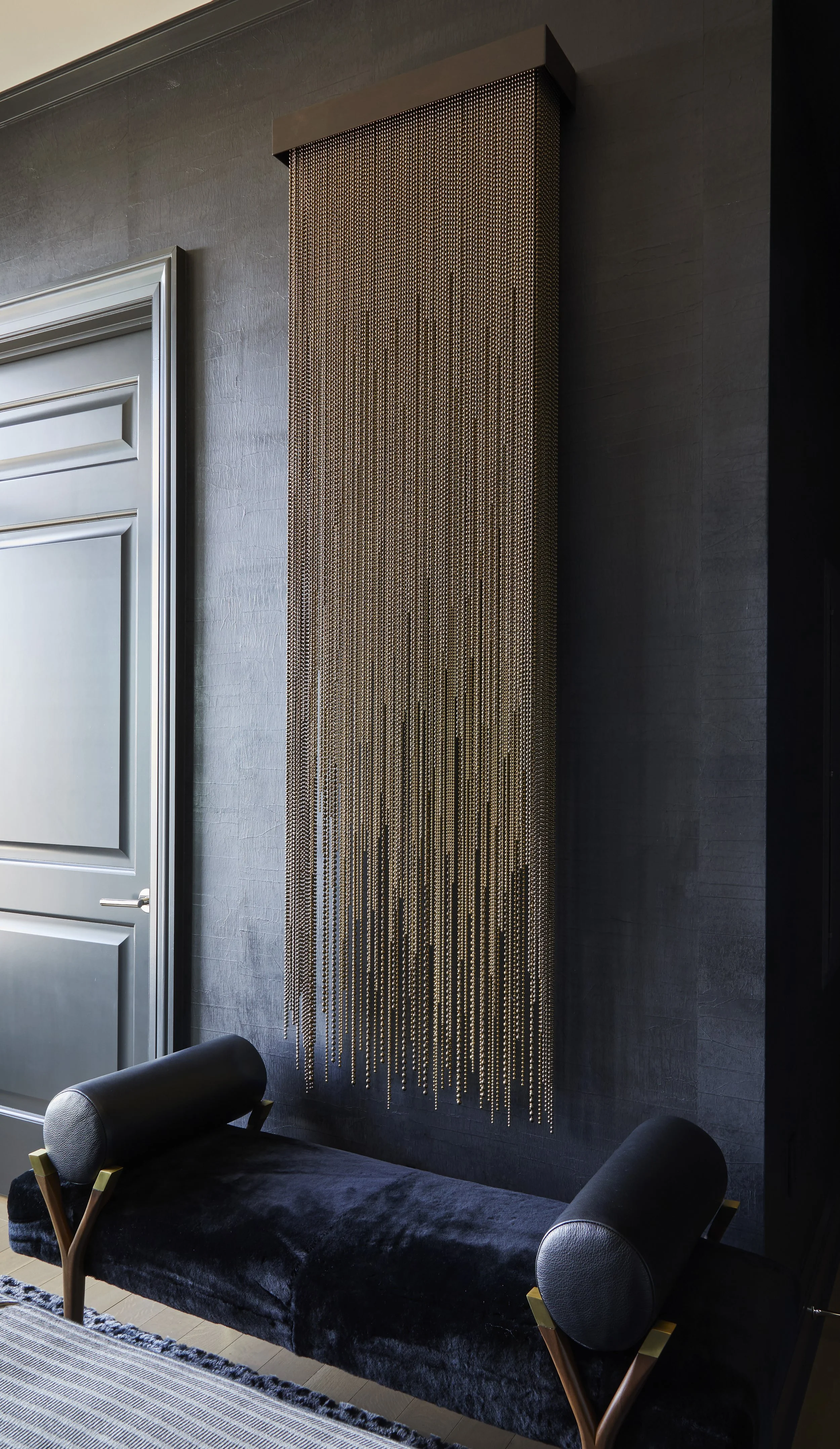 A modern interior space features a dark wall with a vertical metallic beaded curtain hanging on it, a silver-paneled door to the left, and a black velvet upholstered bench with a wooden frame and rounded black armrests below the curtain. Part of a st